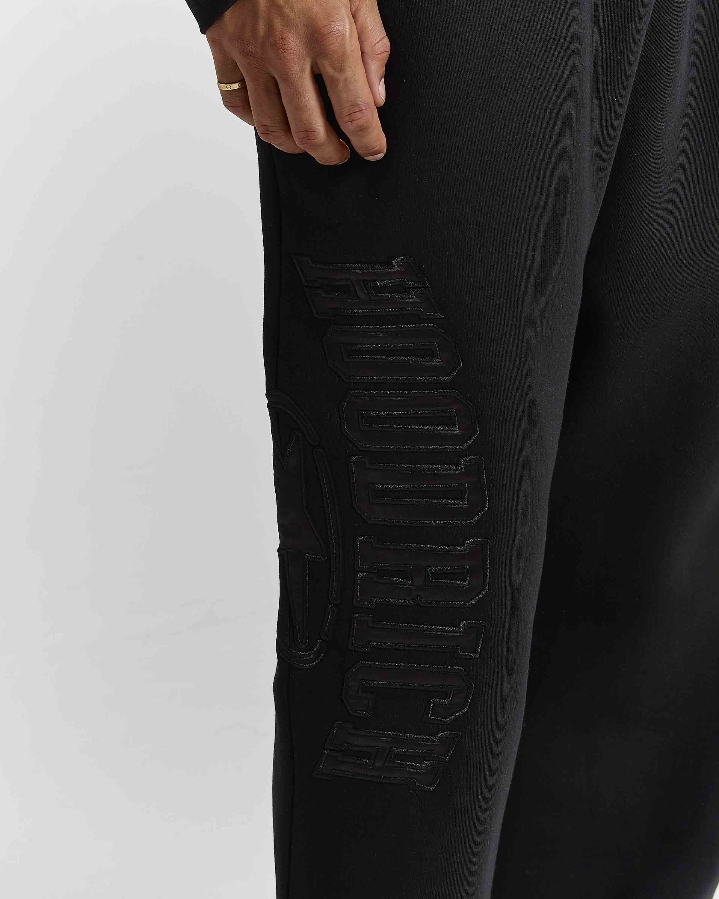 Close-up of a person wearing black sweatpants with large tonal black 'hoodrich' lettering on the side, shown against a light gray studio background.
