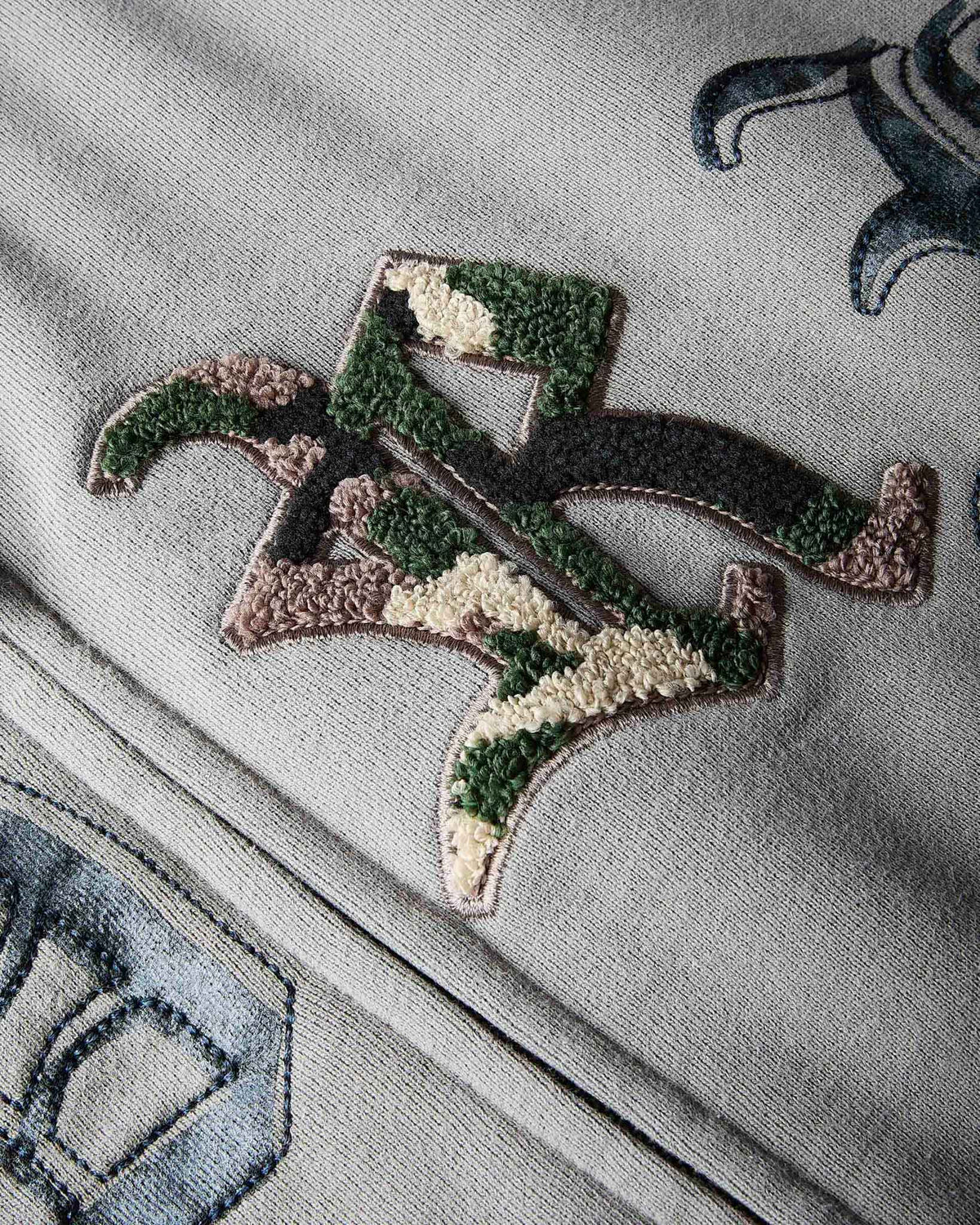 Close-up of a textured embroidered patch featuring a stylized letter 'R' in a green, brown, and beige camouflage pattern, stitched onto a light gray ribbed fabric.