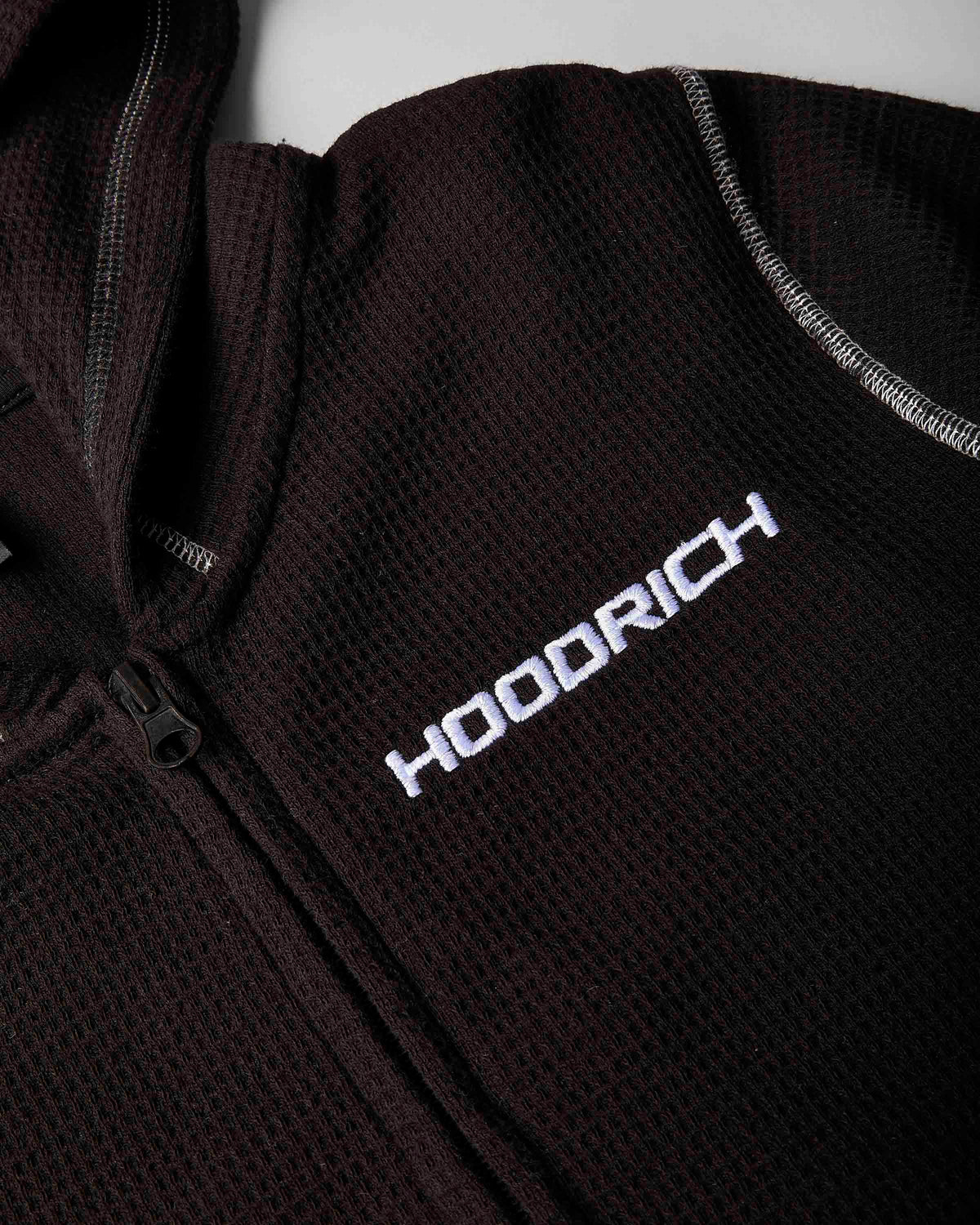 Close-up of a dark brown waffle-knit zip-up jacket with white embroidered 'HOODRICH' branding on the chest.
