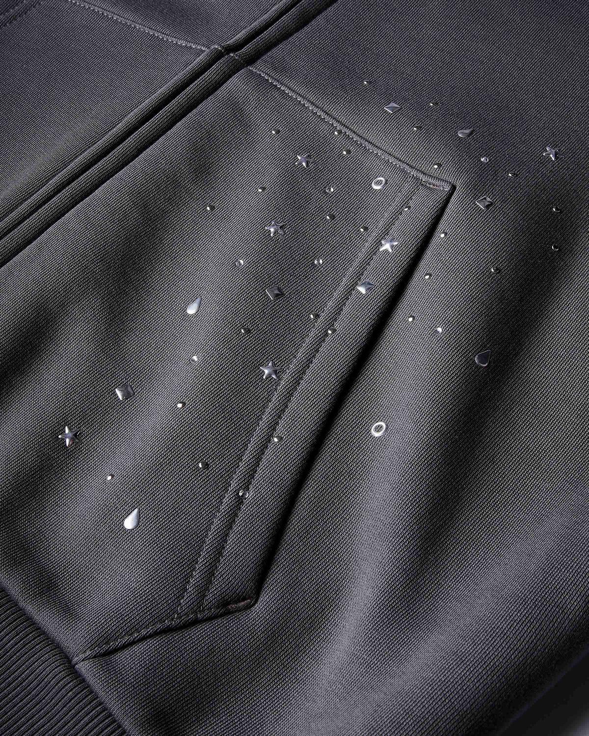 Close-up of a dark charcoal gray fabric surface with a stitched pocket and scattered small reflective silver rhinestone-like studs in star, circle, and teardrop shapes.