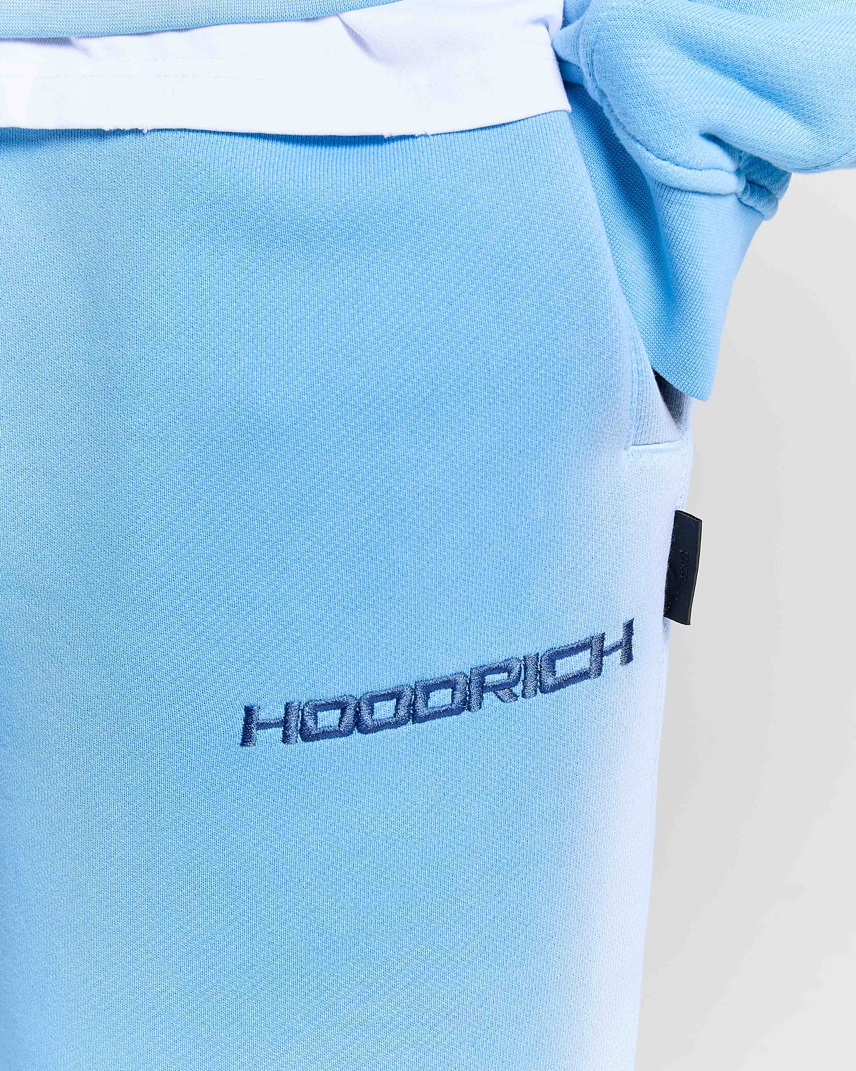 Close-up of the black Hoodrich logo embroidered on the thigh of a model wearing oversized blue joggers.