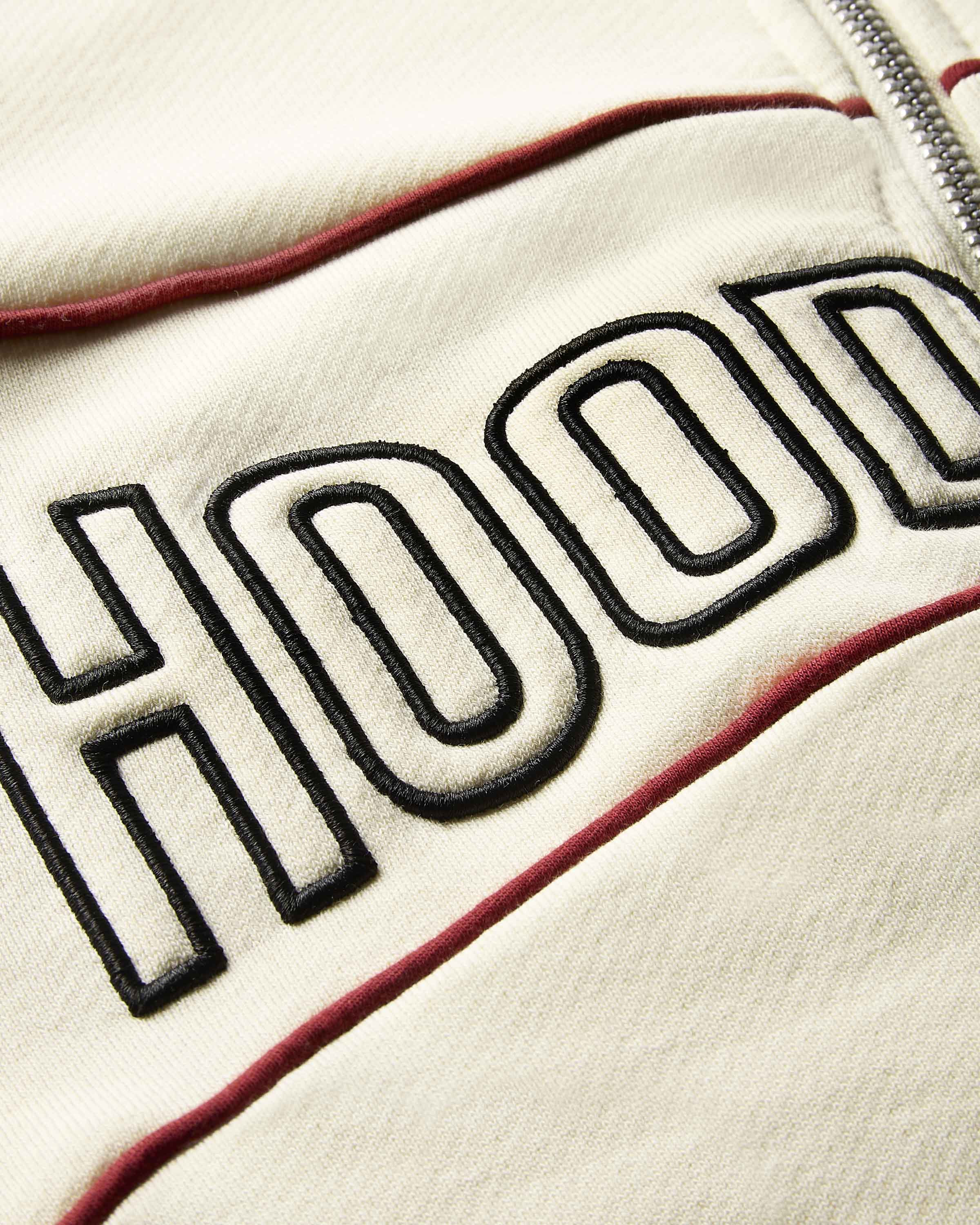 Close-up of a cream-colored zip-up hoodie featuring bold black embroidered lettering and a thin red piping accent.