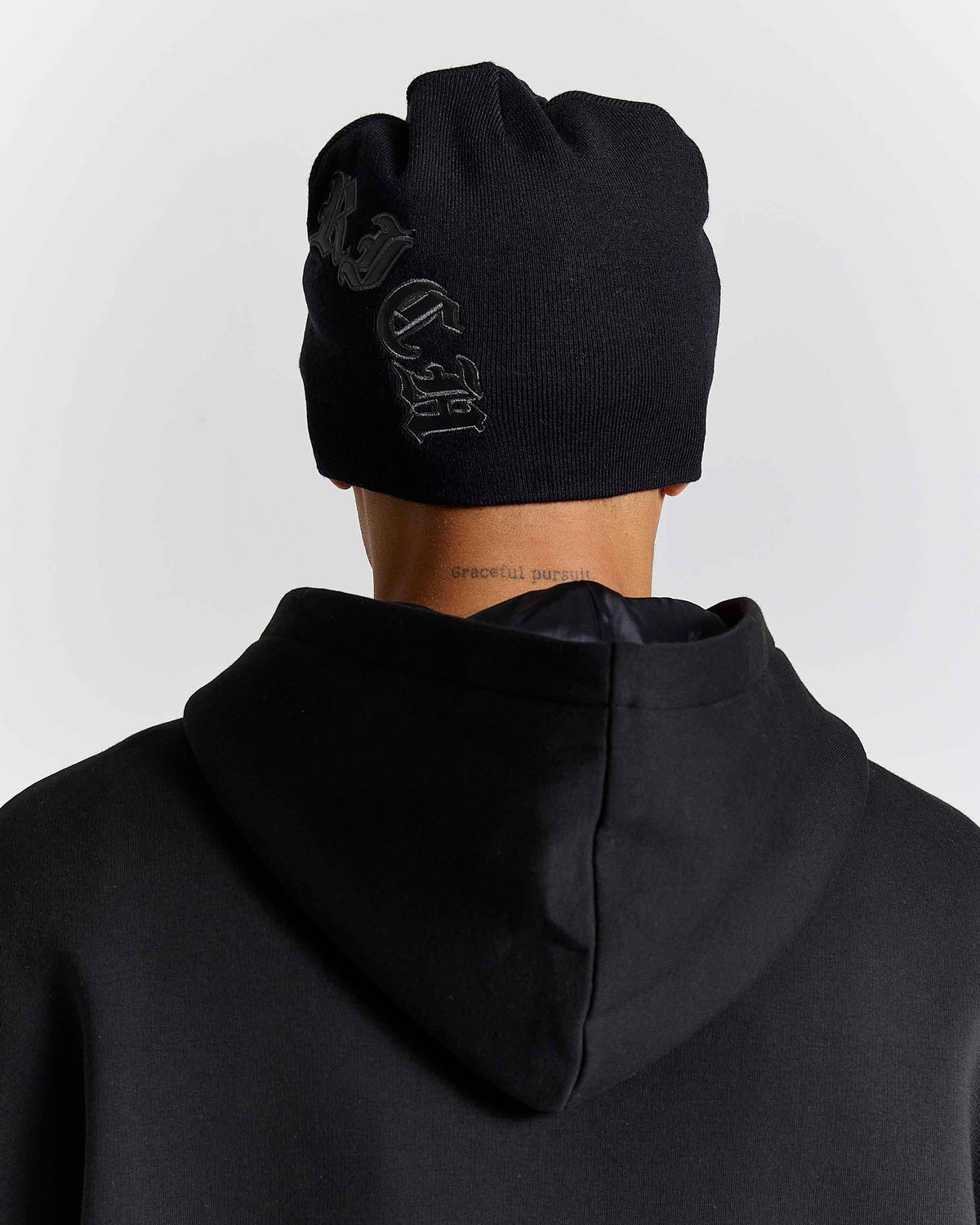 A person shown from the back wearing a black knit beanie with a tonal black embroidered gothic-style logo and a black hoodie against a light gray studio background.