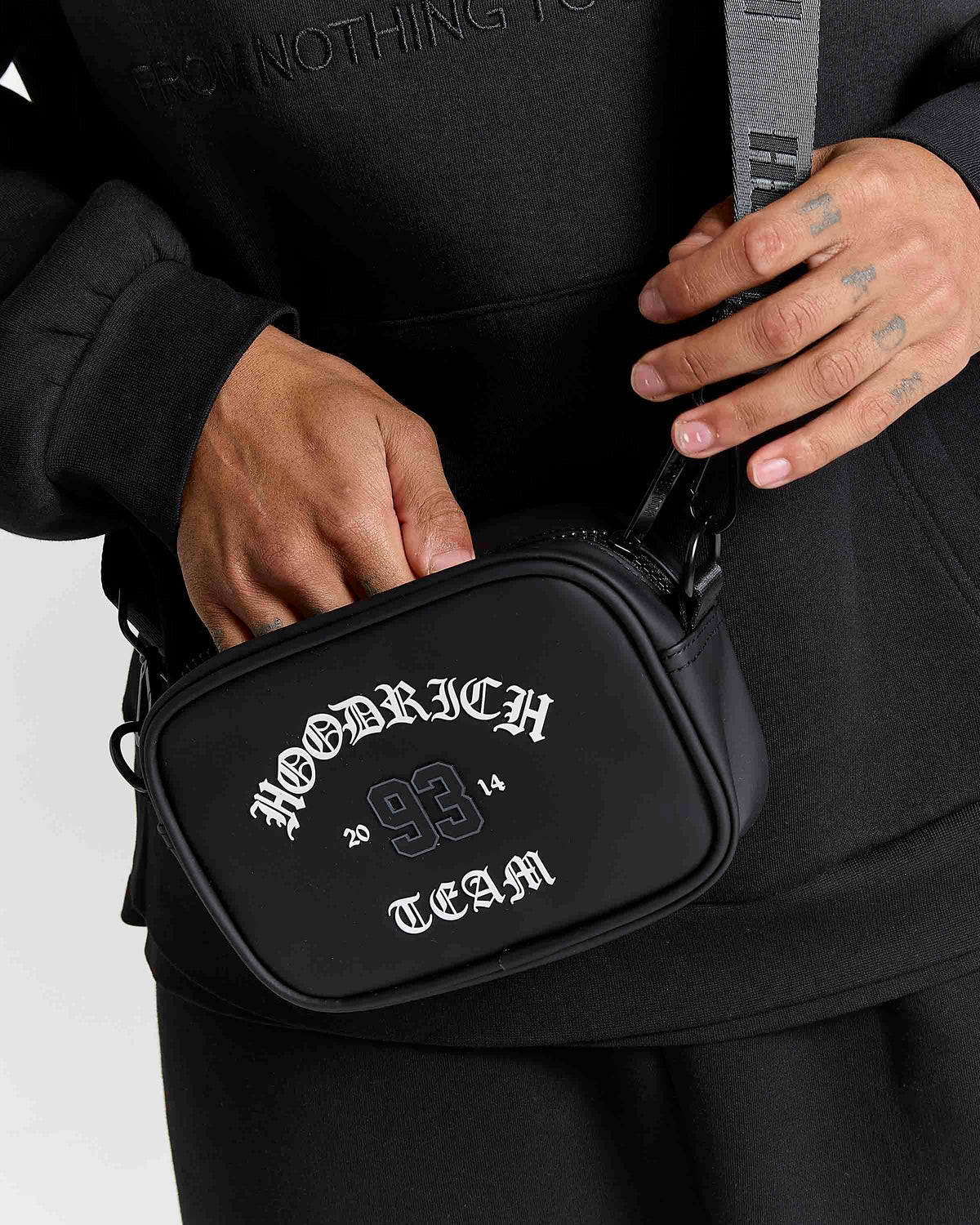 A black crossbody bag with white gothic lettering reading 'HOODRICH TEAM' being worn by a person in a black hoodie. The person's hands and tattooed arms are visible holding the bag and strap.