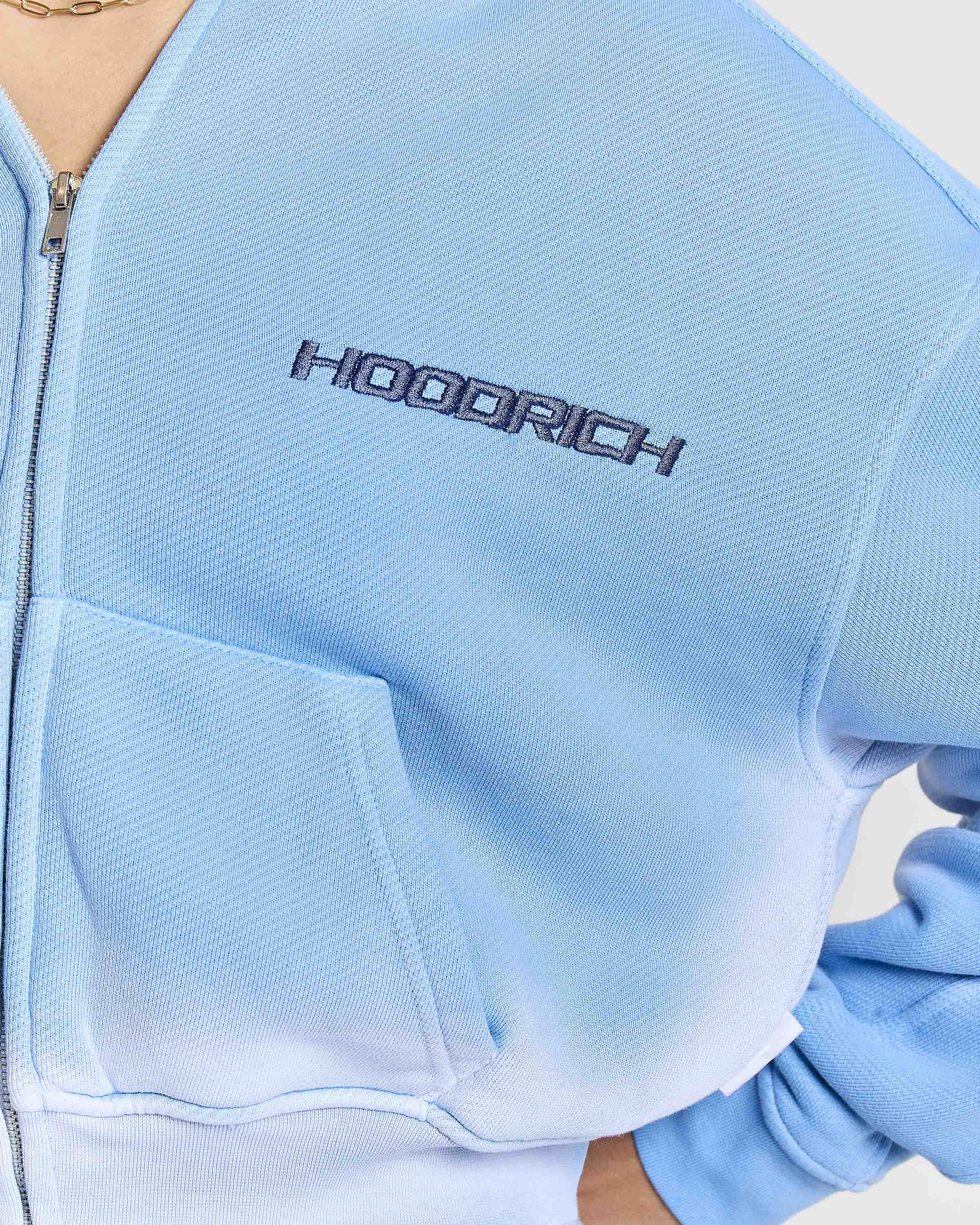 Close-up of the Hoodrich logo embroidered on a model wearing a blue ombre zip-up cropped hoodie.