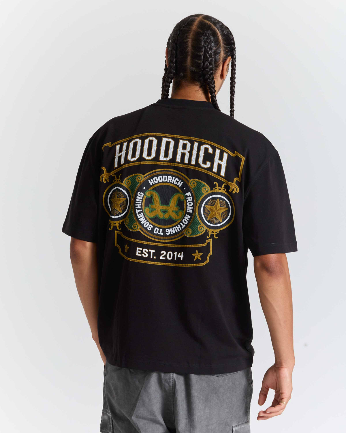 A man viewed from the back wearing a black HOODRICH t-shirt with a large gold and white graphic print on the back, paired with gray cargo pants against a light gray studio background.