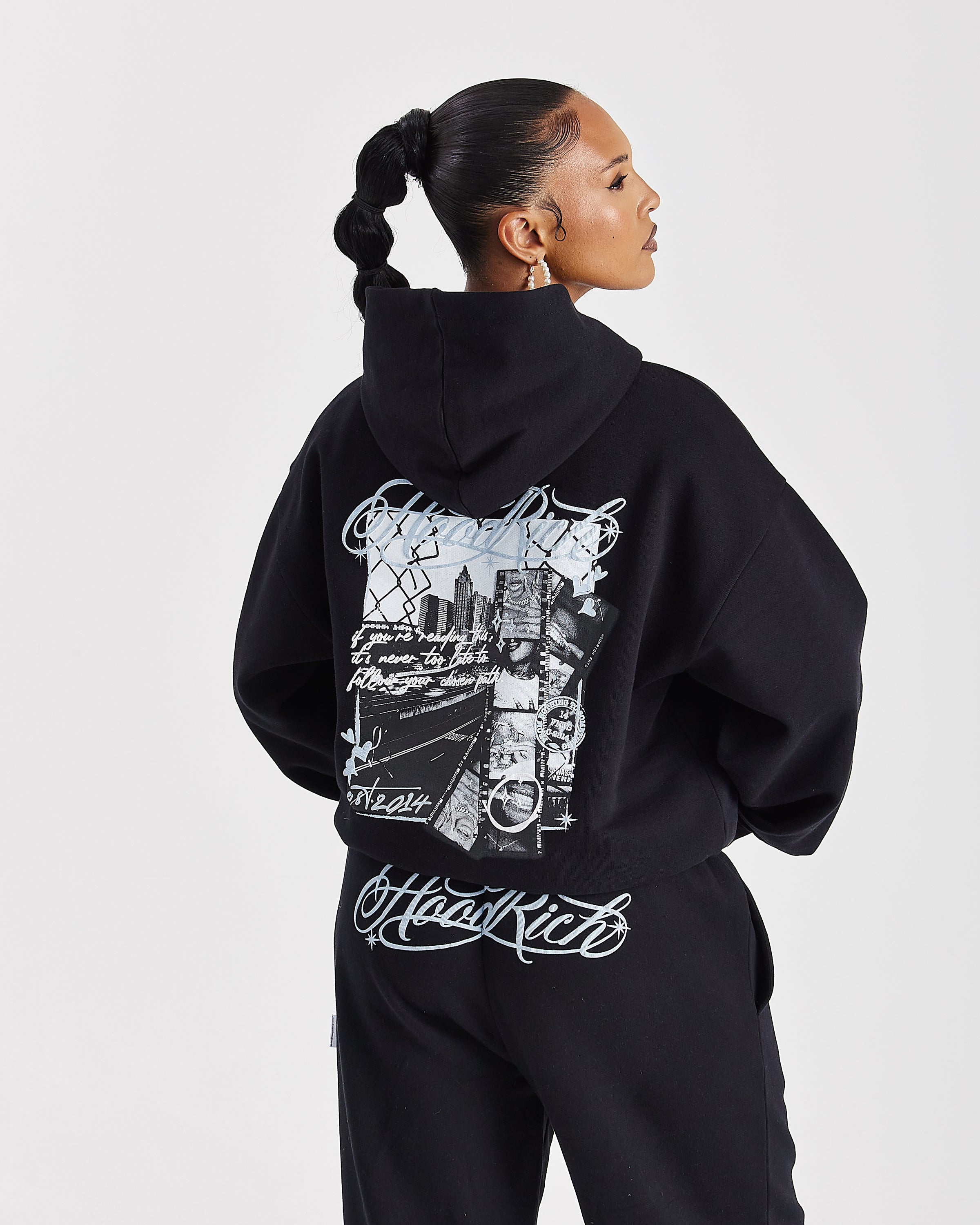 A model shown from the back wearing a black hoodie and matching sweatpants set, featuring a large white graphic print on the back of the hoodie and 'Hoodrich' written in white script near the hem.