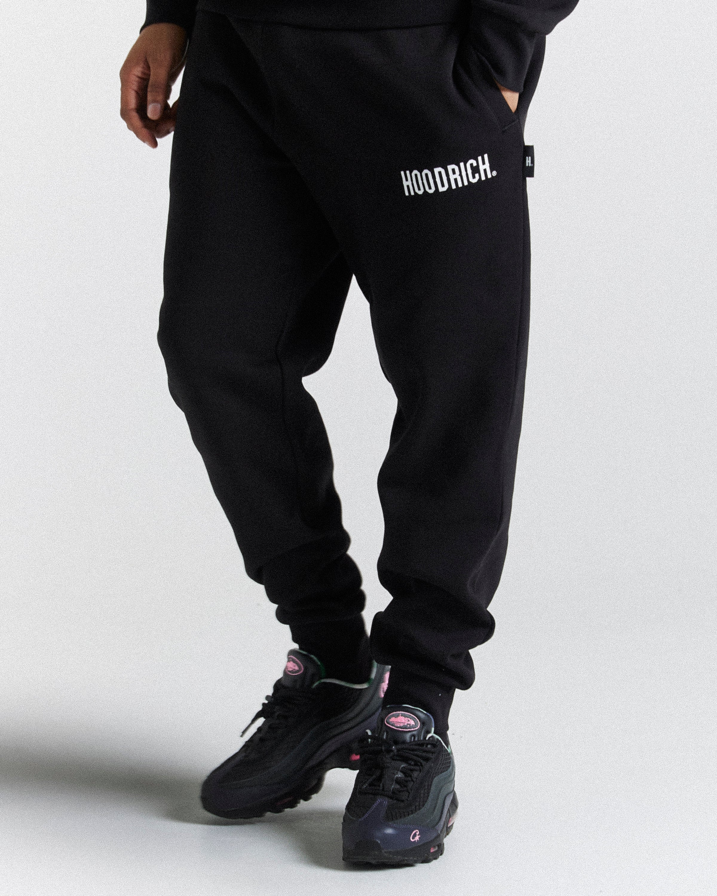 Black sweatpants with 'HOODRICH.' printed in white on the upper left thigh, shown on a model wearing black sneakers against a light gray studio background.