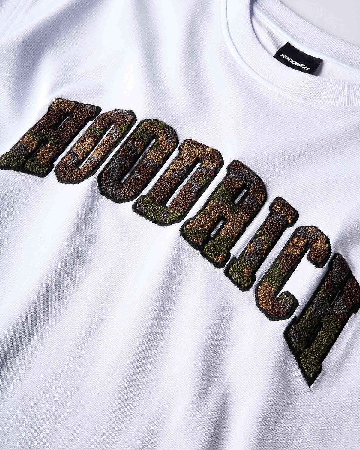 Close-up of a white crewneck t-shirt featuring a large arched graphic across the chest in textured, metallic-look letters with a camouflage pattern of brown, green, and gold. The brand name 'HOODRICH' is clearly spelled out.