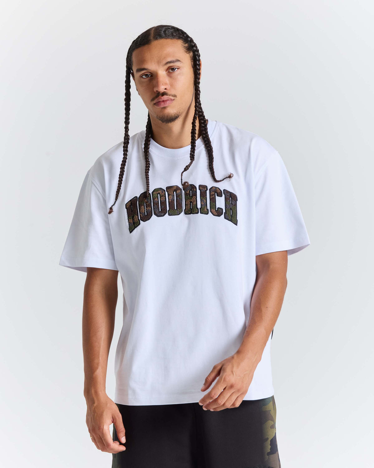 A man with long braids wearing a white Woodrich t-shirt with a dark camouflage-style arched Hoodrich  logo across the chest, paired with black pants, against a light gray studio background.