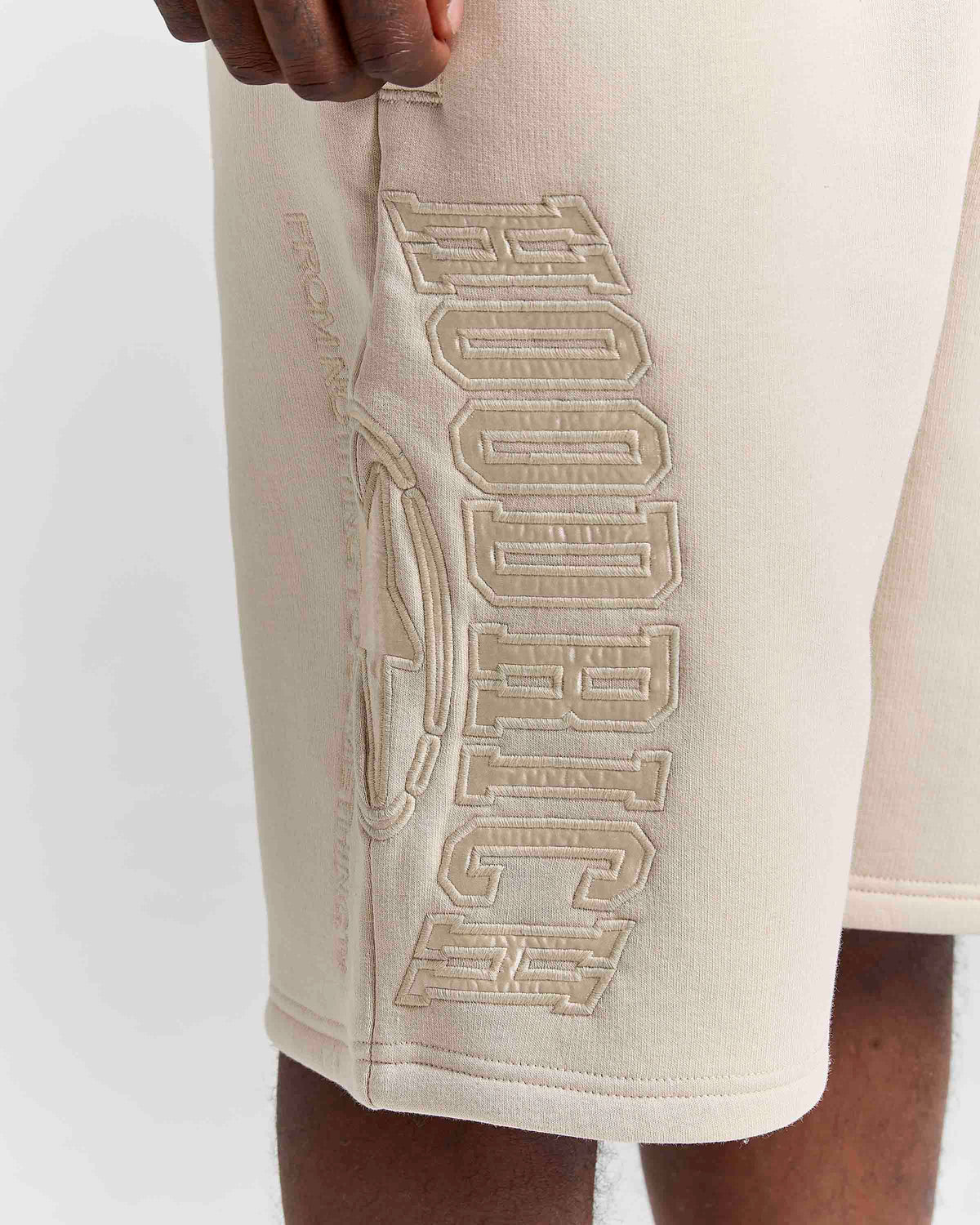 Close-up of a person wearing cream-colored athletic shorts with large tonal embroidered lettering on the side.