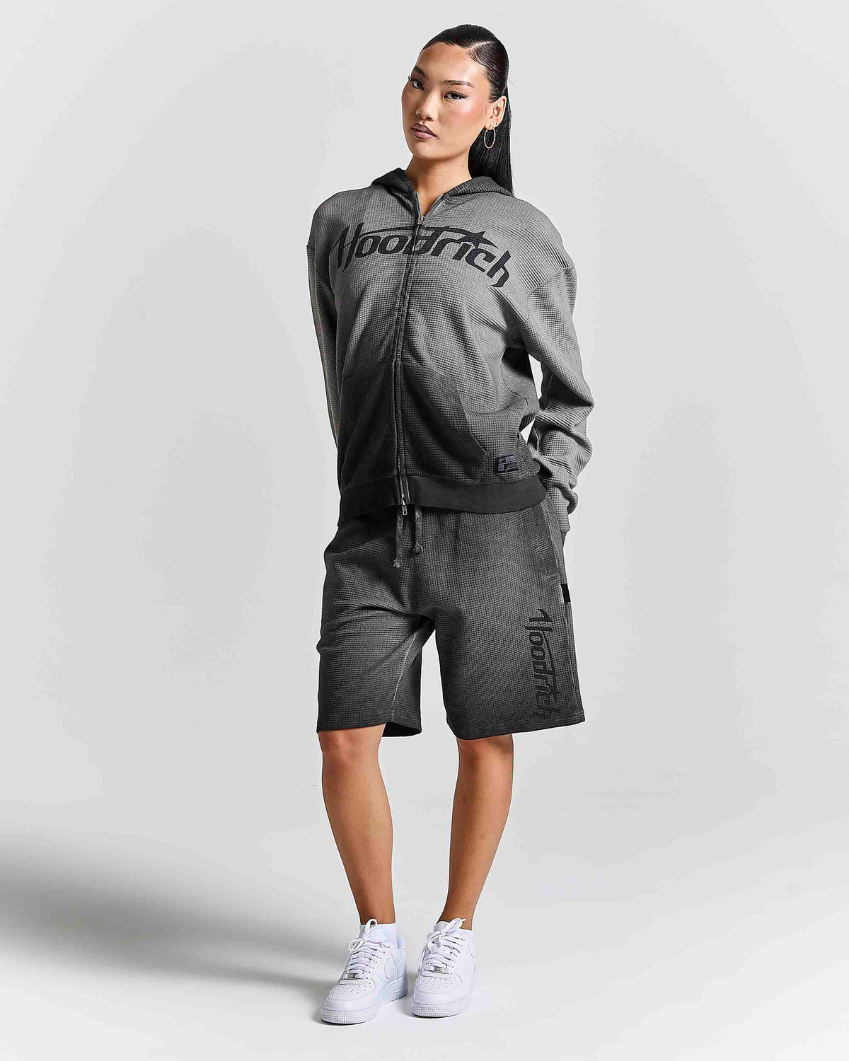 A woman modeling a dark gray two-piece tracksuit featuring a zip-up hoodie with 'Hoodrich' printed across the chest and matching shorts with 'AlexRich' printed down the leg, paired with white sneakers against a light gray studio background.