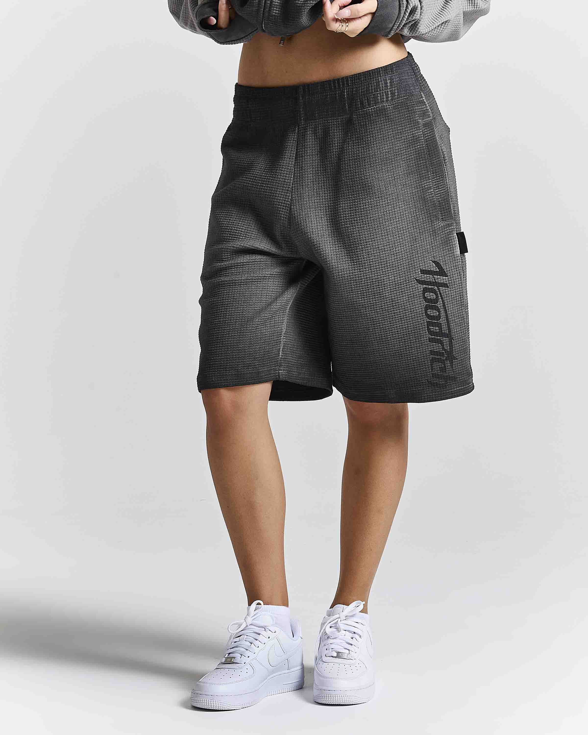 A person wearing dark gray textured athletic shorts with a vertical black 'Hoodrich' logo on the side, paired with white sneakers and a gray cropped jacket, shown from the waist down against a light gray studio background.
