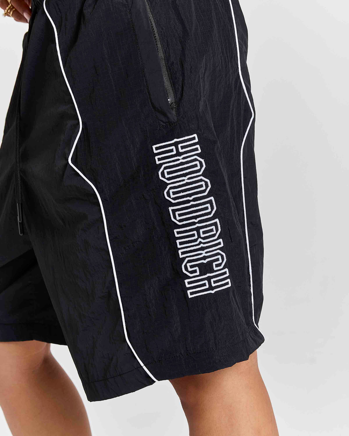 Close-up of black athletic shorts with white piping and a large white outlined vertical 'HOODRICH' logo on the side, worn by a model against a light gray background.