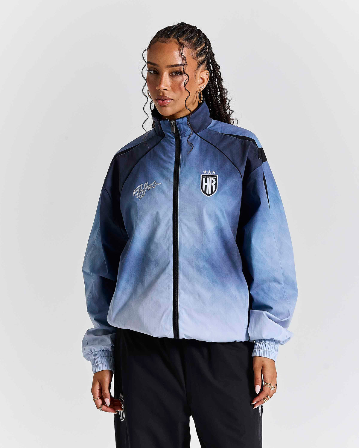 A model wearing a blue and navy gradient zip-up jacket with a black and white 'HR' shield logo on the chest, paired with black pants, against a light gray studio background.'