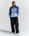 A man modeling a light blue and navy gradient zip-up jacket with black track pants featuring light blue star details, set against a plain light gray studio background.
