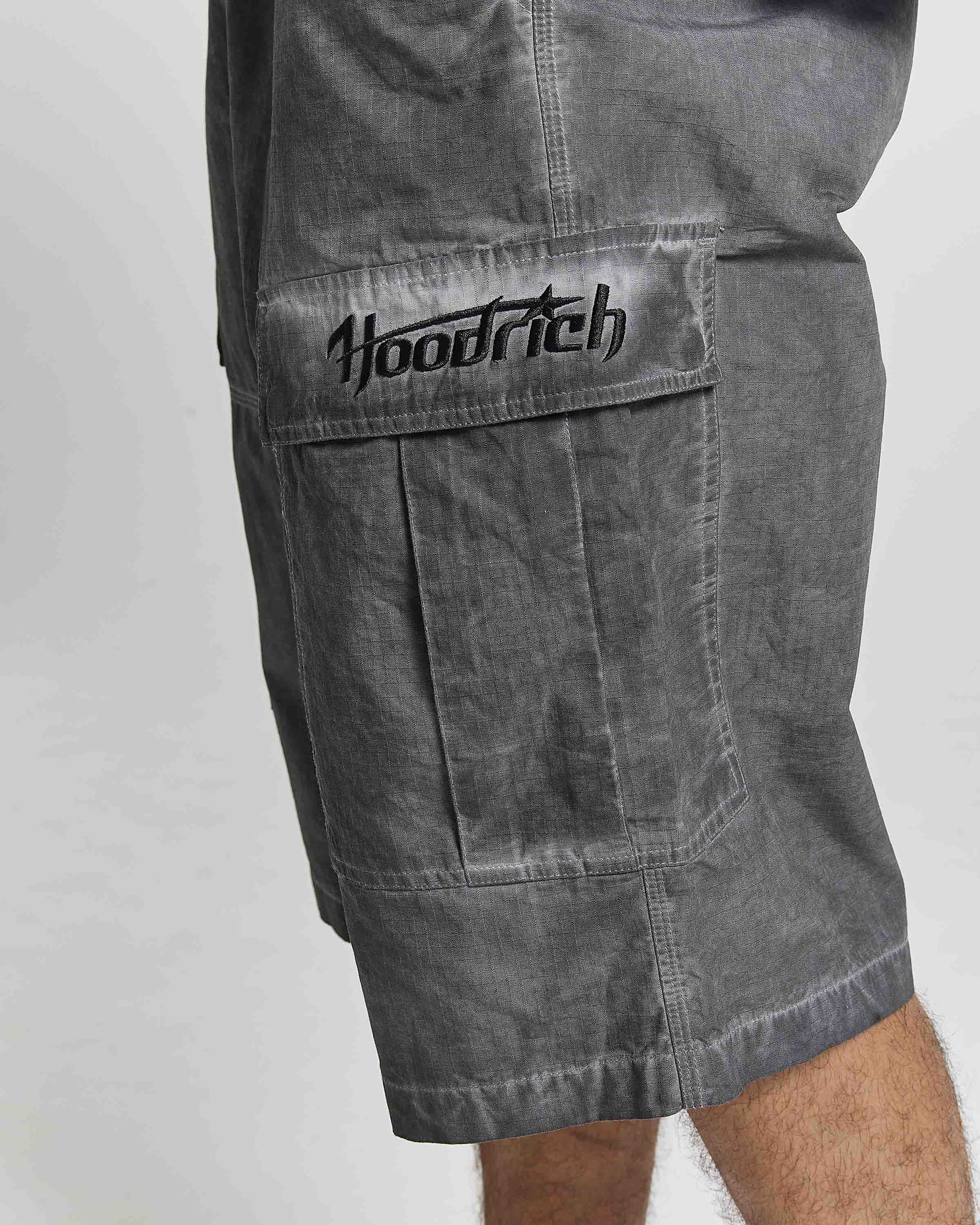 Close-up of a person wearing gray washed cargo shorts with a large front flap pocket and black embroidered 'Hoodrich' logo on the pocket, against a light gray background.