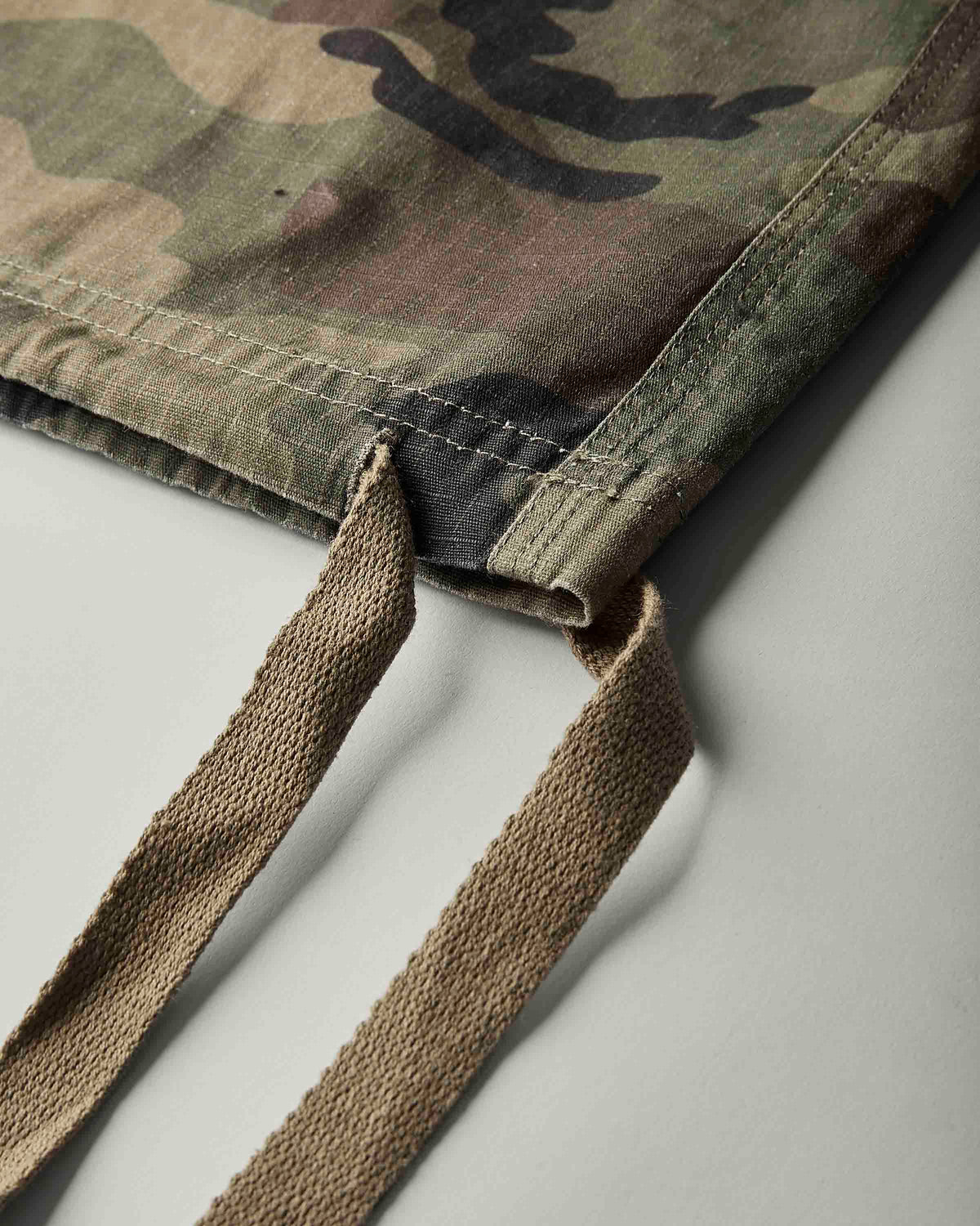 Close-up of a camouflage fabric bag corner with tan woven drawstring straps, photographed on a light gray background.