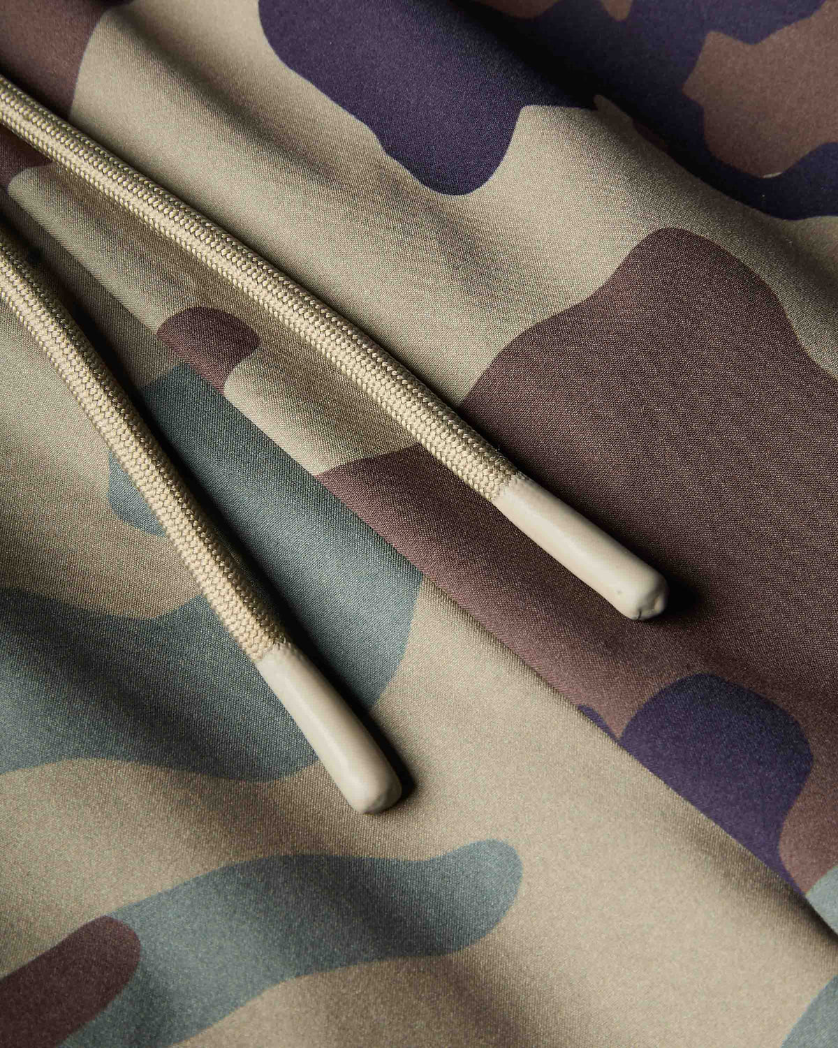 Close-up of a camouflage fabric product with beige rope drawstrings and cream tips, showing a pattern of beige, brown, gray, and muted purple.