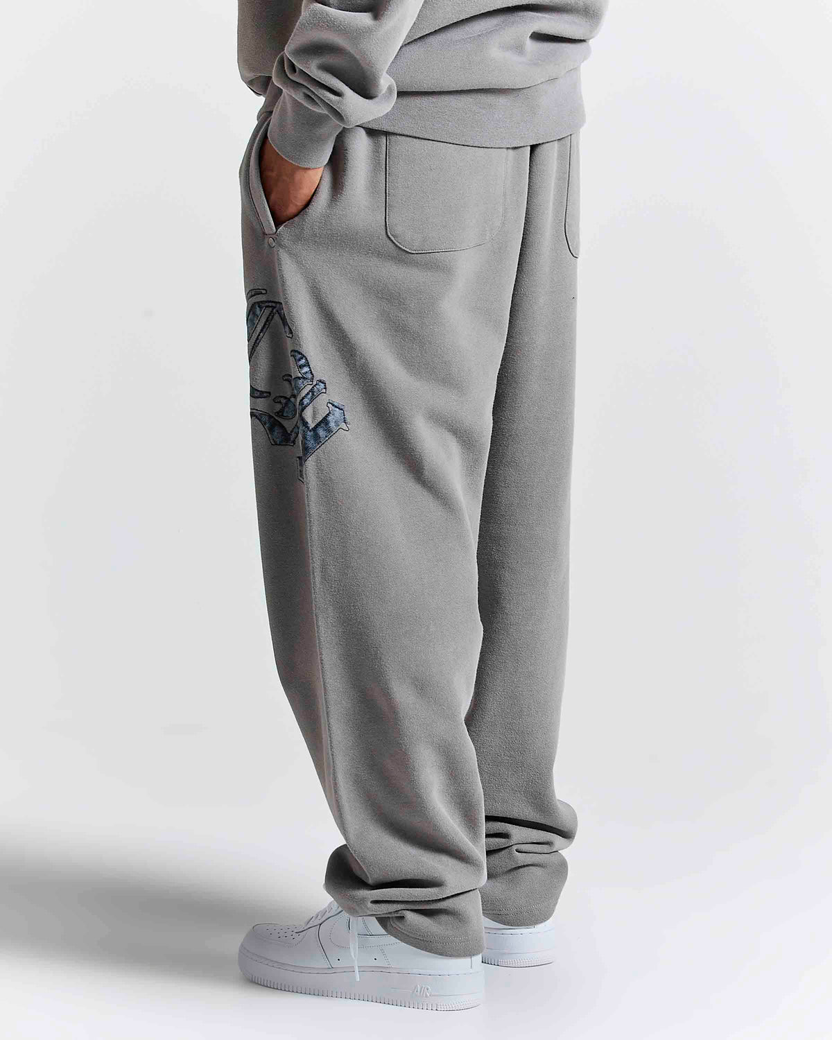 A person wearing light gray sweatpants with a blue graphic print on the left leg, paired with a matching gray sweatshirt and white sneakers, shown from the waist down against a light gray studio background.