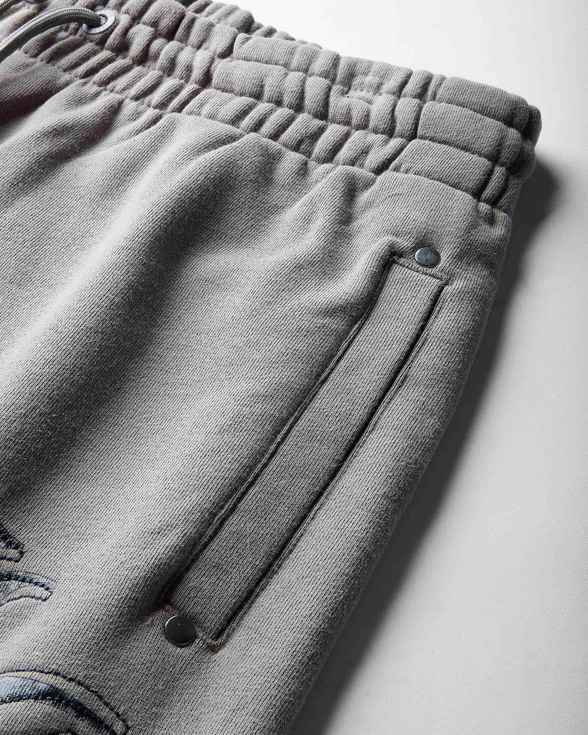 Close-up of gray fleece sweatpants showing the elastic waistband, a side pocket with metal snap closure, and part of a tonal embroidered logo on a light gray background.