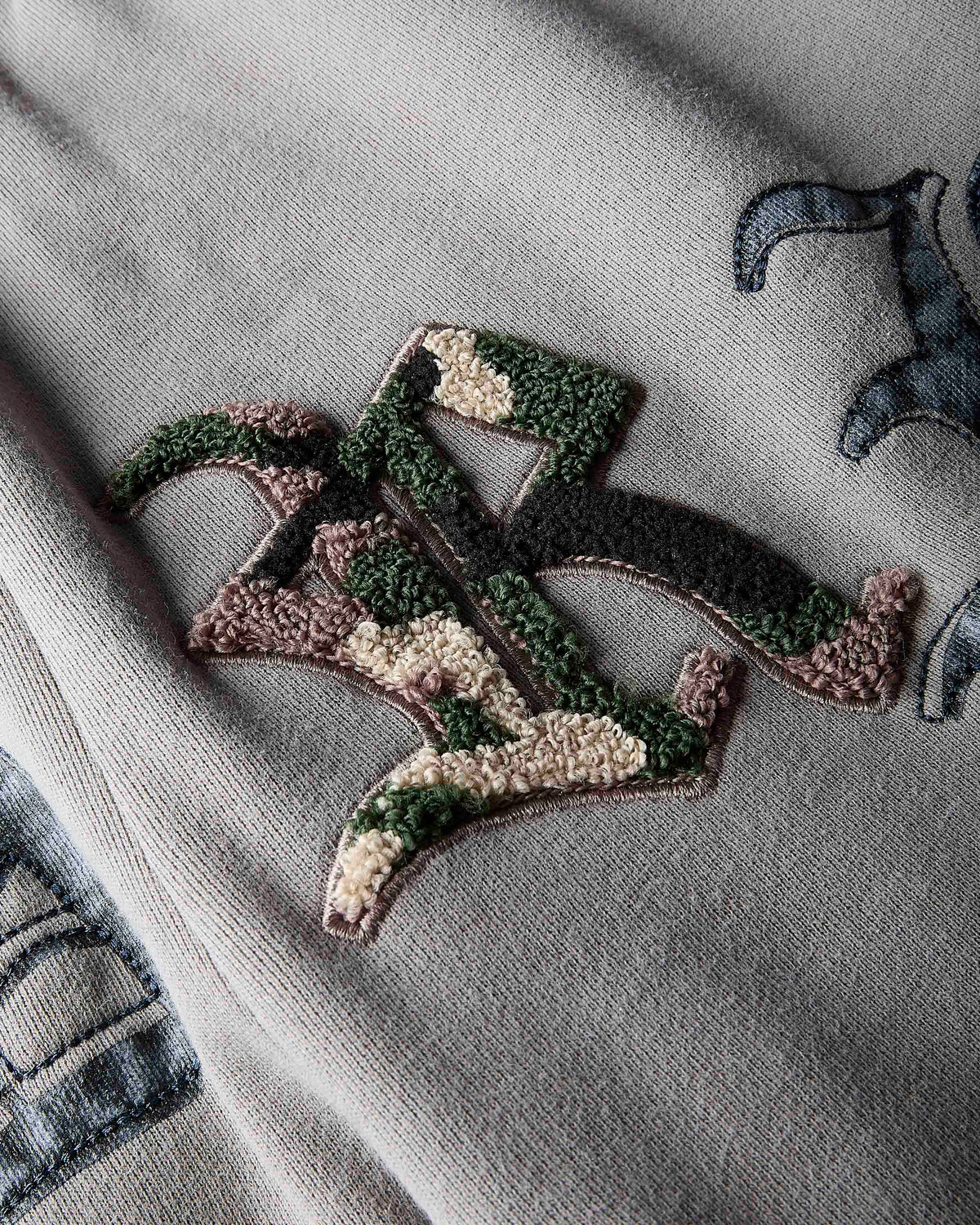 A close-up of a textured gray knit garment featuring a large, stylized embroidered patch in green, beige, and brown camouflage-like thread, with other partial embroidered patches visible around the edges.