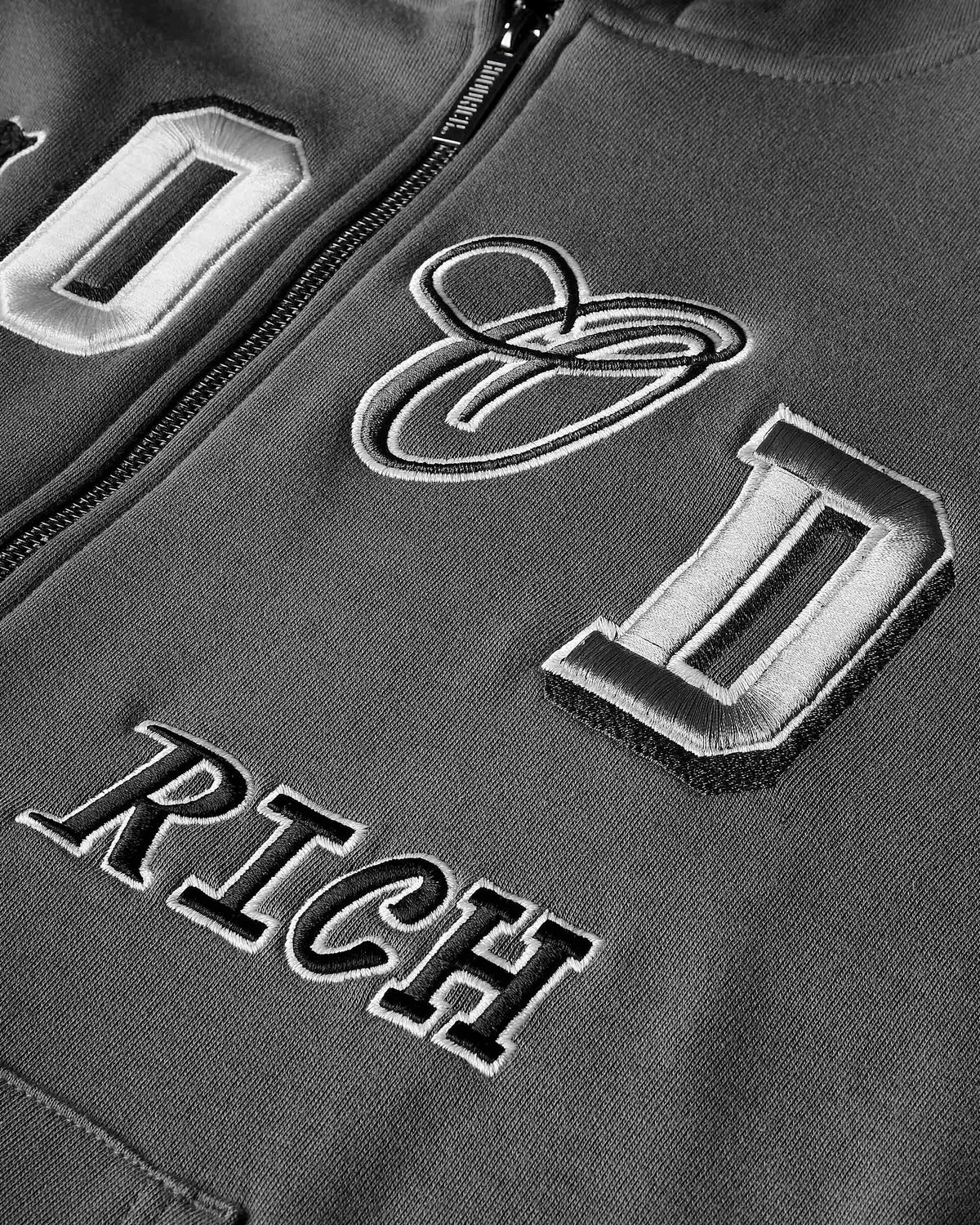 Close-up of a dark gray zip-up jacket featuring large embroidered varsity-style letters and the word 'HOODRICH' in black and white.