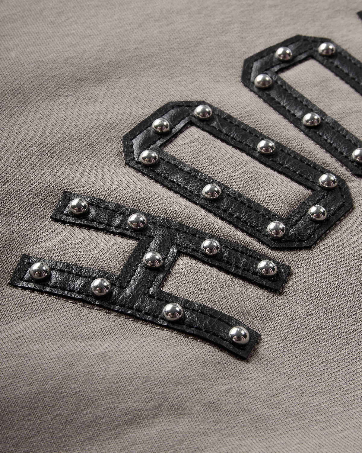 Close-up of a taupe knit fabric featuring large black faux-leather embroidered letters adorned with silver-tone metal studs.