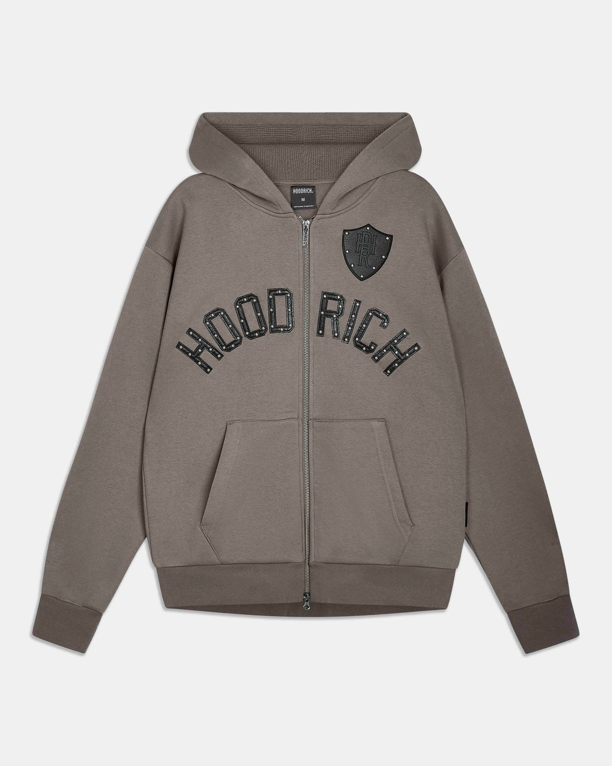 A taupe-brown zip-up hoodie with 'HOODRICH' printed in black arched lettering across the chest and a black shield emblem on the left chest, shown against a light gray background.