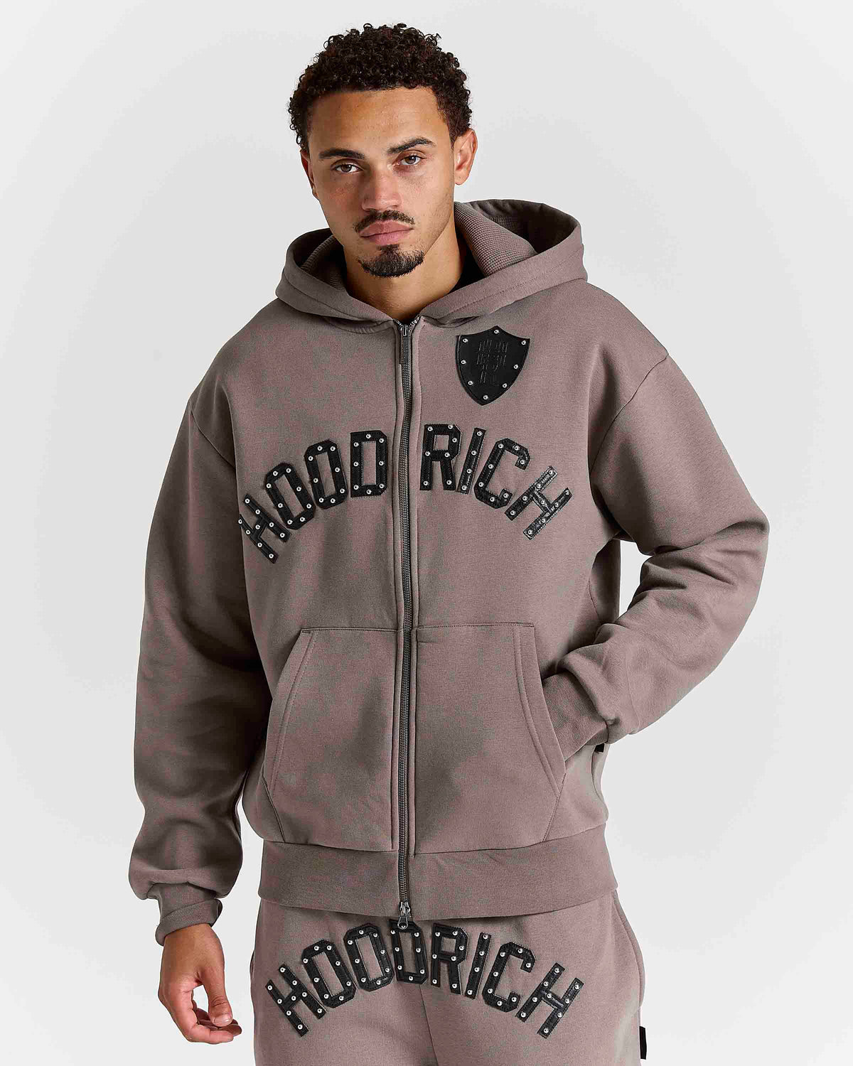 Displays a model wearing the Brown 'Hoodrich' branded men's Varso Studs Full Tracksuit 