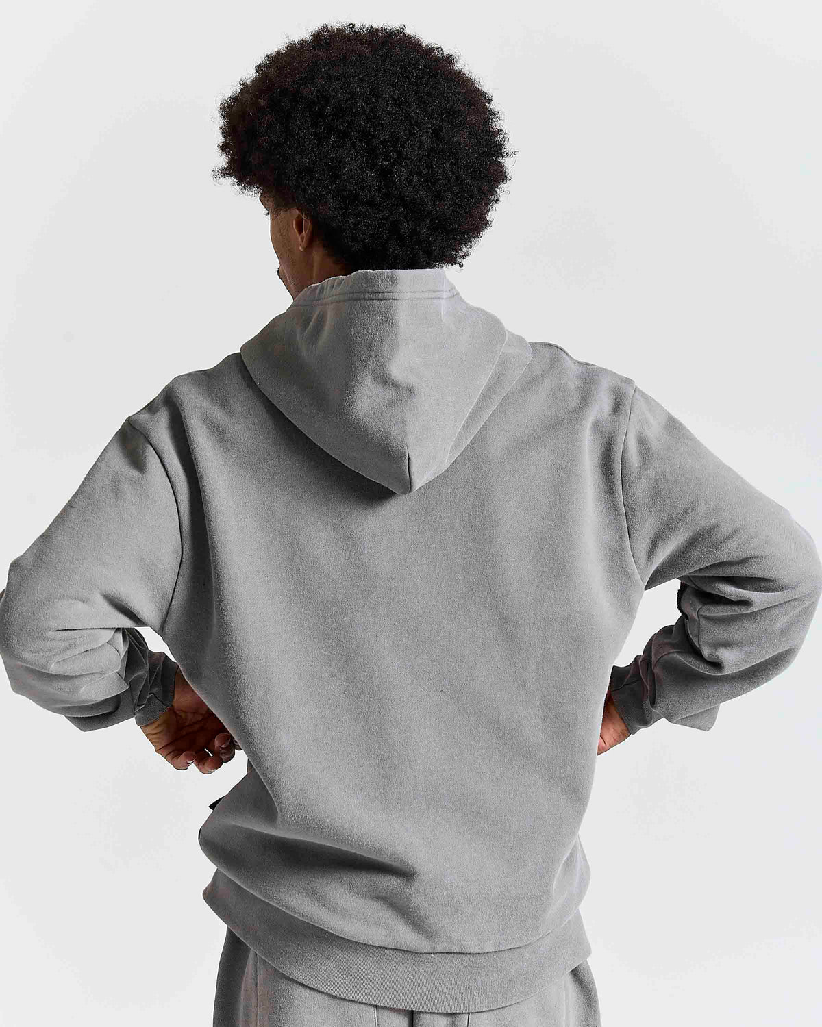 A person shown from the back wearing a light gray pullover hoodie and matching sweatpants against a plain light gray studio background.