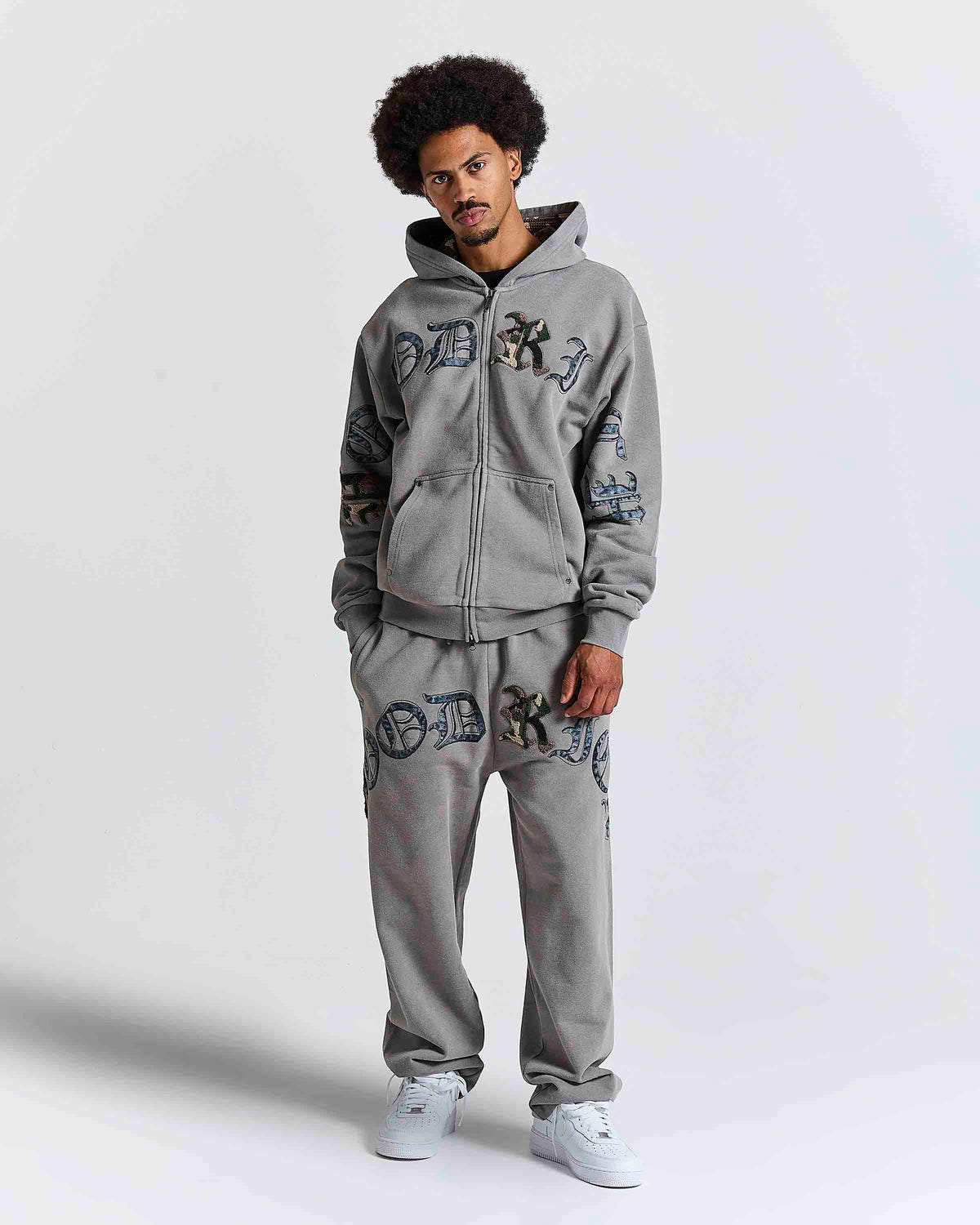 A man modeling a gray zip-up hoodie and matching sweatpants set with blue and black gothic-style letter embroidery, set against a plain light gray studio background.