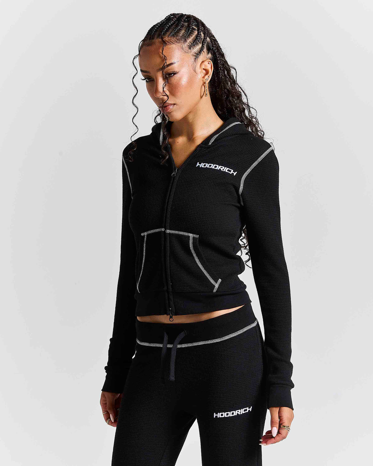 A woman modeling a black zip-up hoodie and matching black joggers with white contrast stitching and white HOODRICH logos, photographed against a light gray studio background.