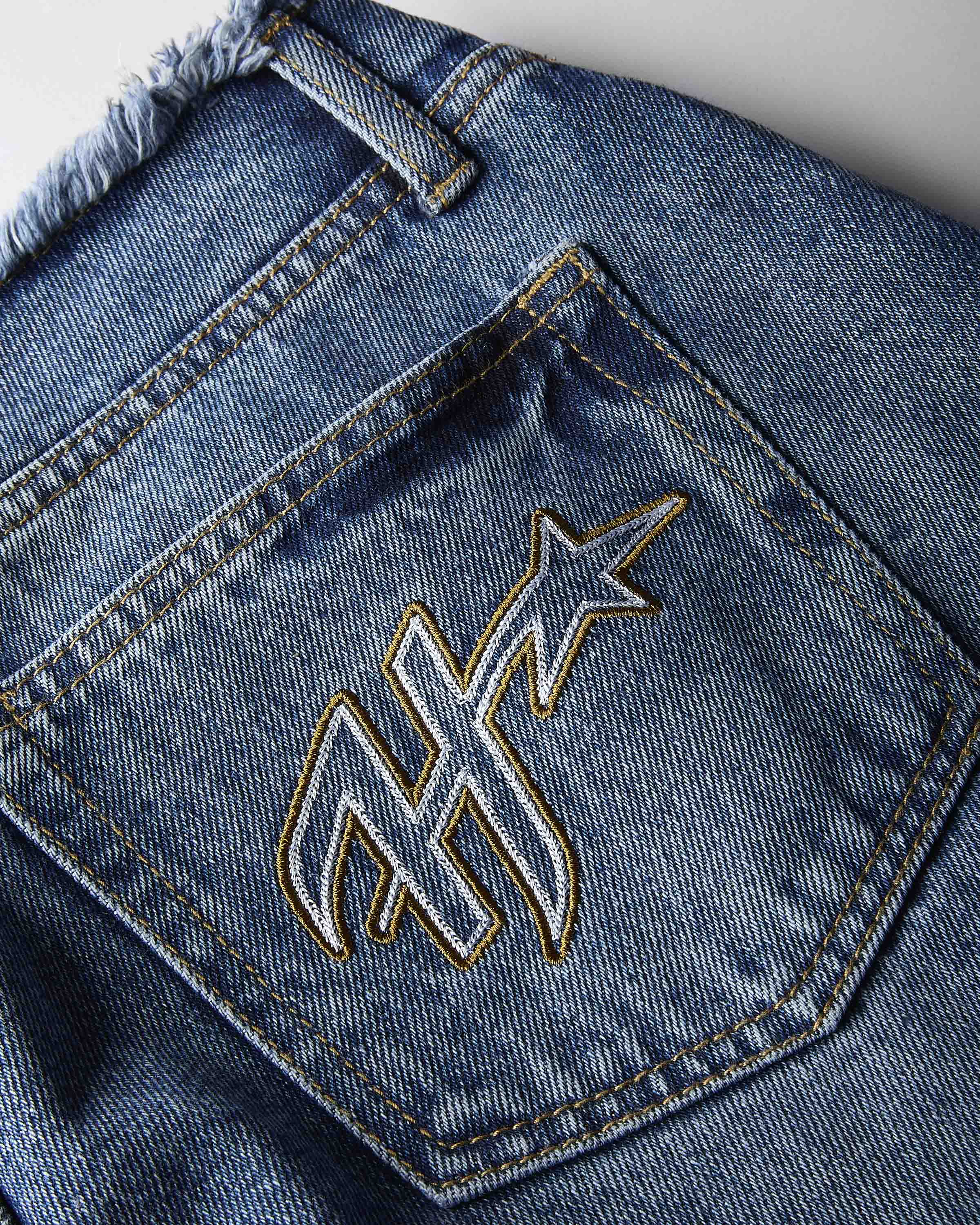 Close-up of the back pocket of blue denim jeans with a large embroidered monogram logo outlined in gold thread.