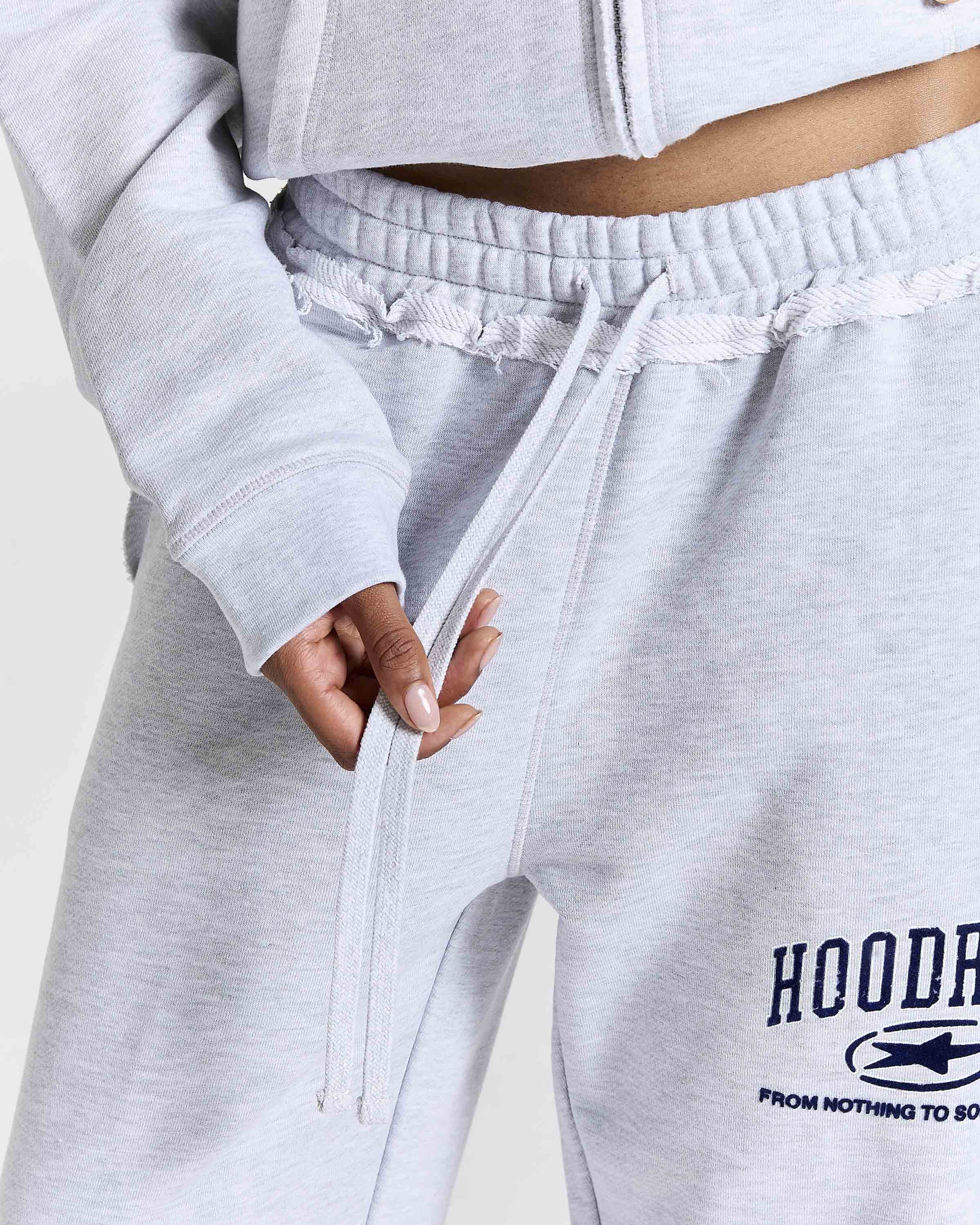 Close-up of a person wearing light heather gray sweatpants with a drawstring waist and a navy graphic print on the left leg that reads 'HOODRICH'