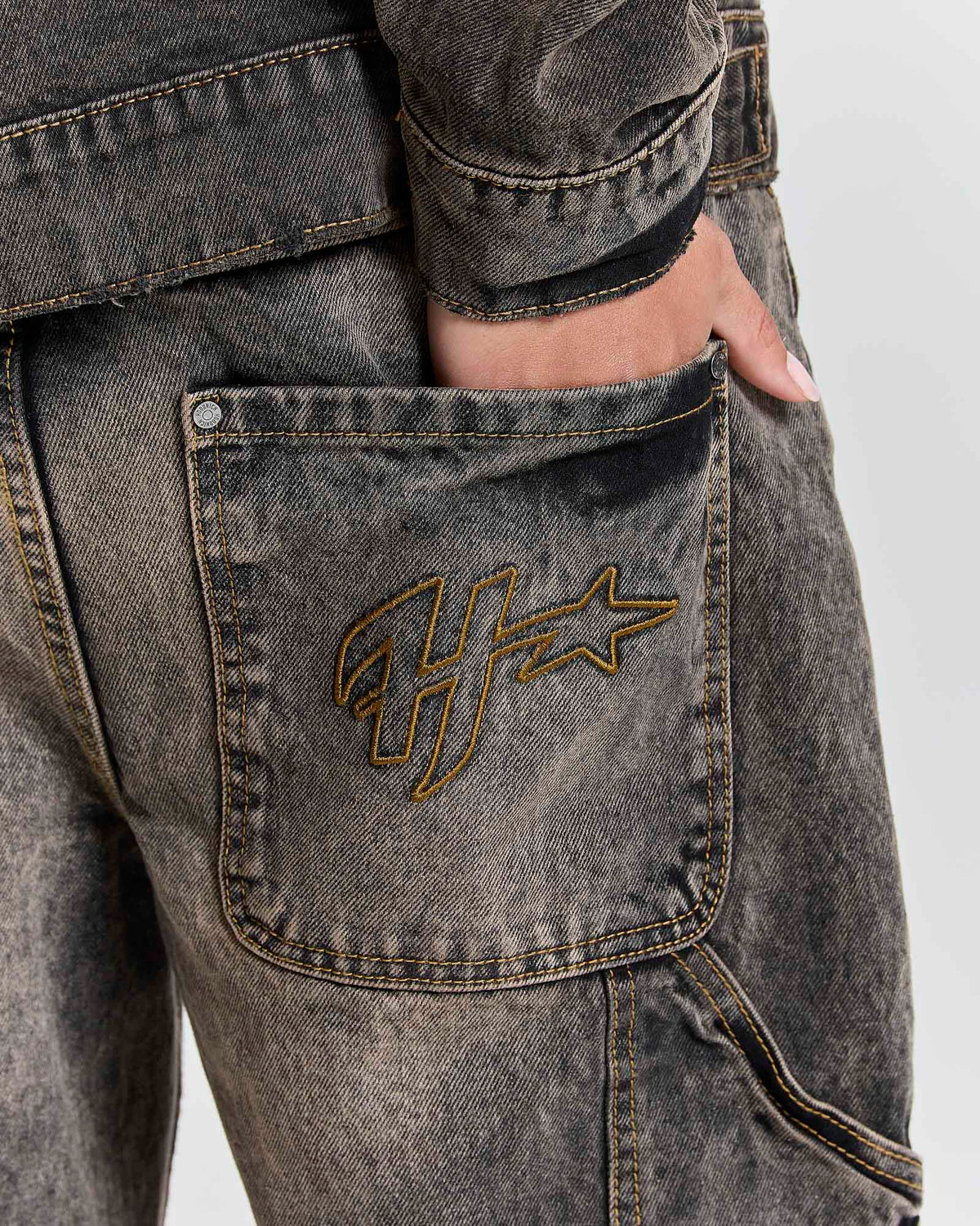 Close-up of a person wearing washed charcoal gray denim jeans with a gold embroidered Hoodrich  logo on the back pocket, showing a hand in the pocket.