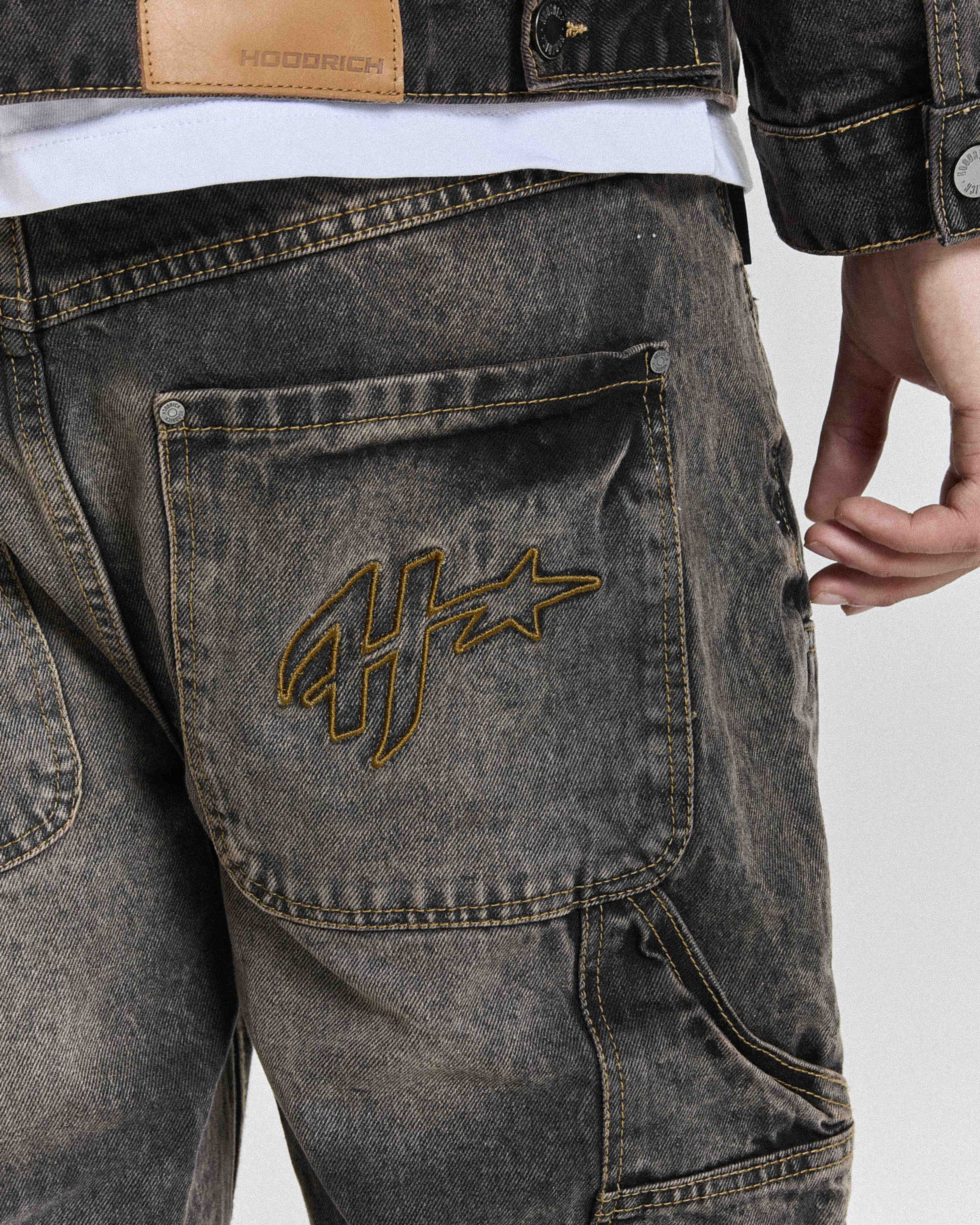 Close-up of the Hoodrich logo embroidered on the pocket of the regular fit black rust washed denim jorts worn by a model.