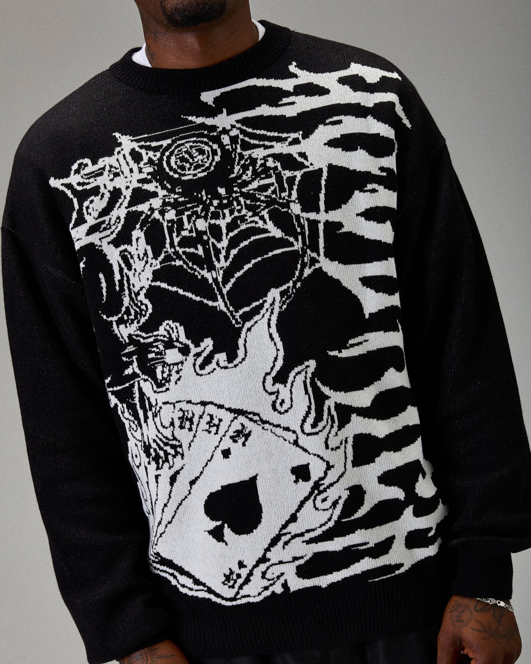Black sweater with white graphic design featuring a spiderweb, card, and other elements on a gray background.