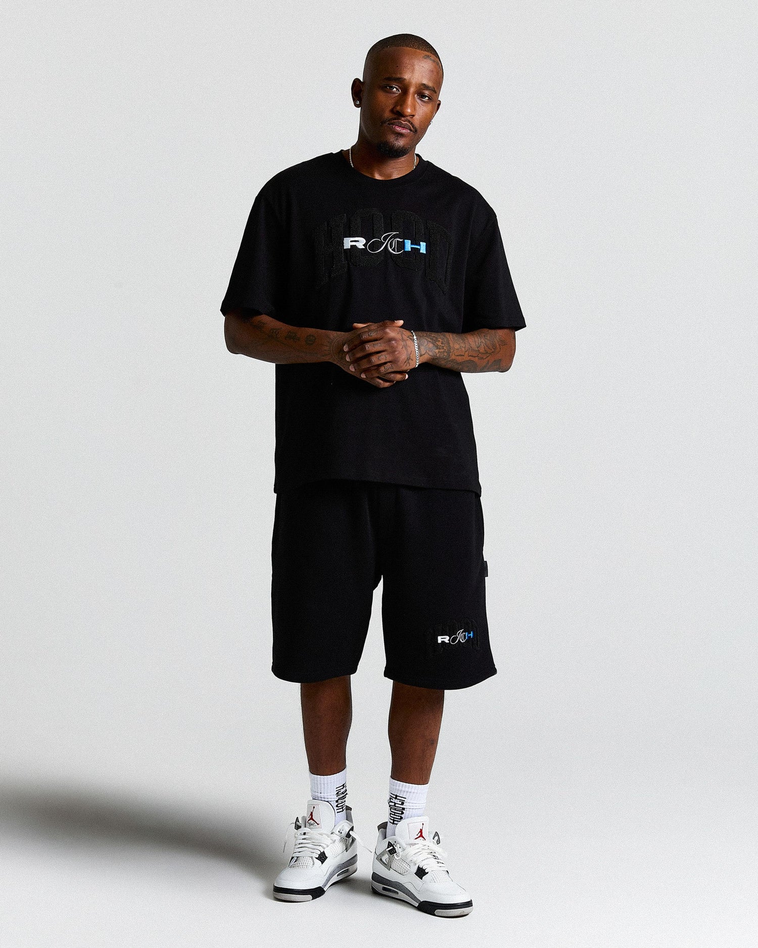 Man wearing a black t-shirt and shorts with logos, standing against a white background