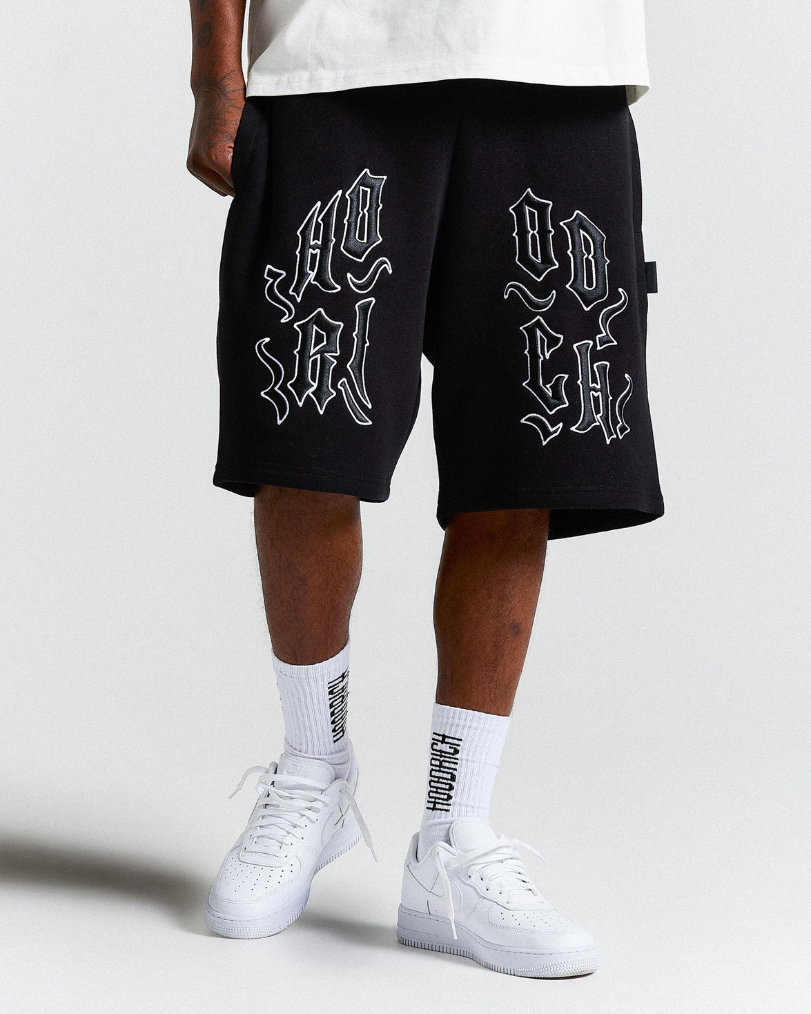 Person wearing black shorts with white text and white sneakers on a light gray background