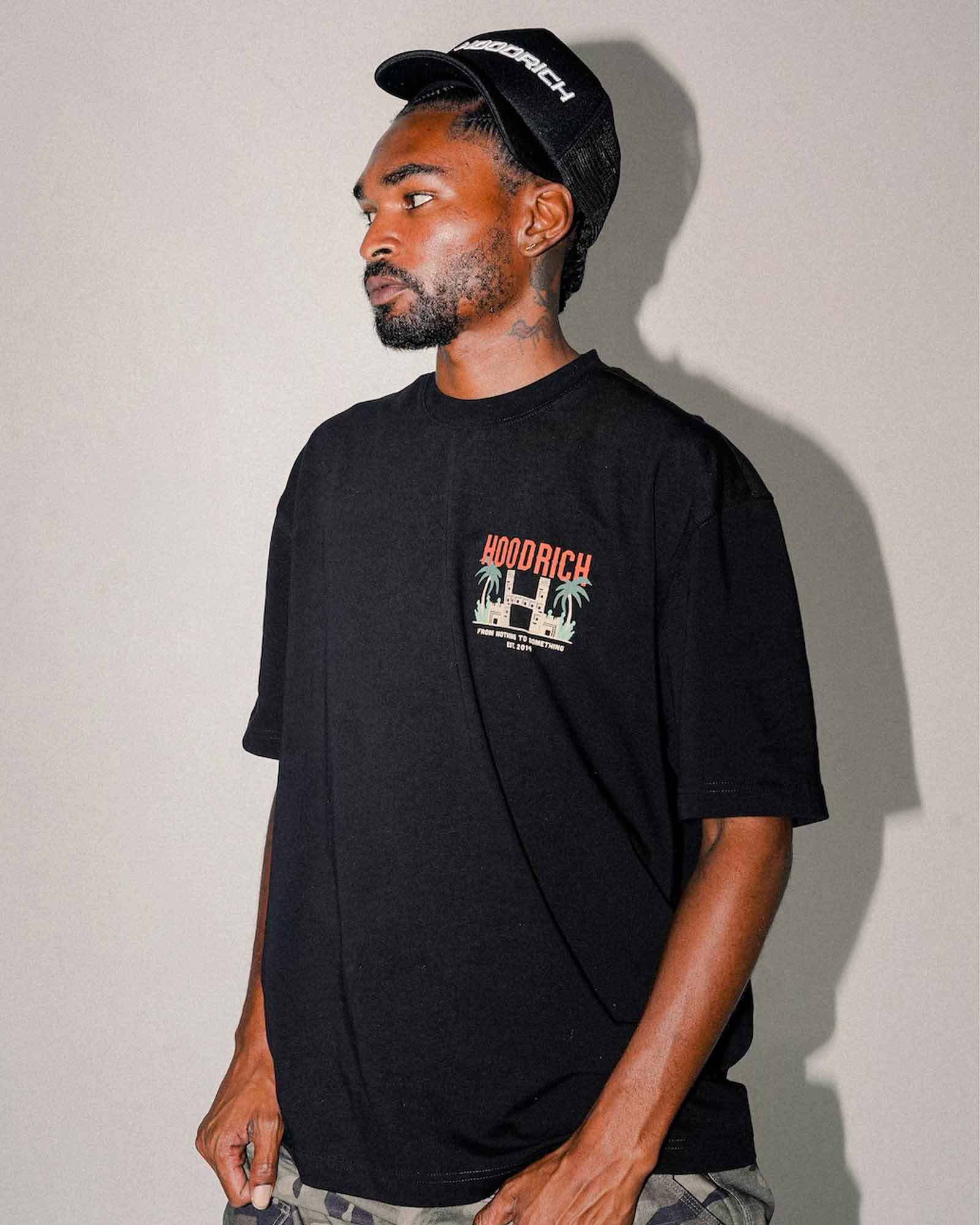 A man wearing a black Hoodrich t-shirt with a graphic on the chest, a black Hoodrich cap, and camouflage pants, standing against a plain gray wall.