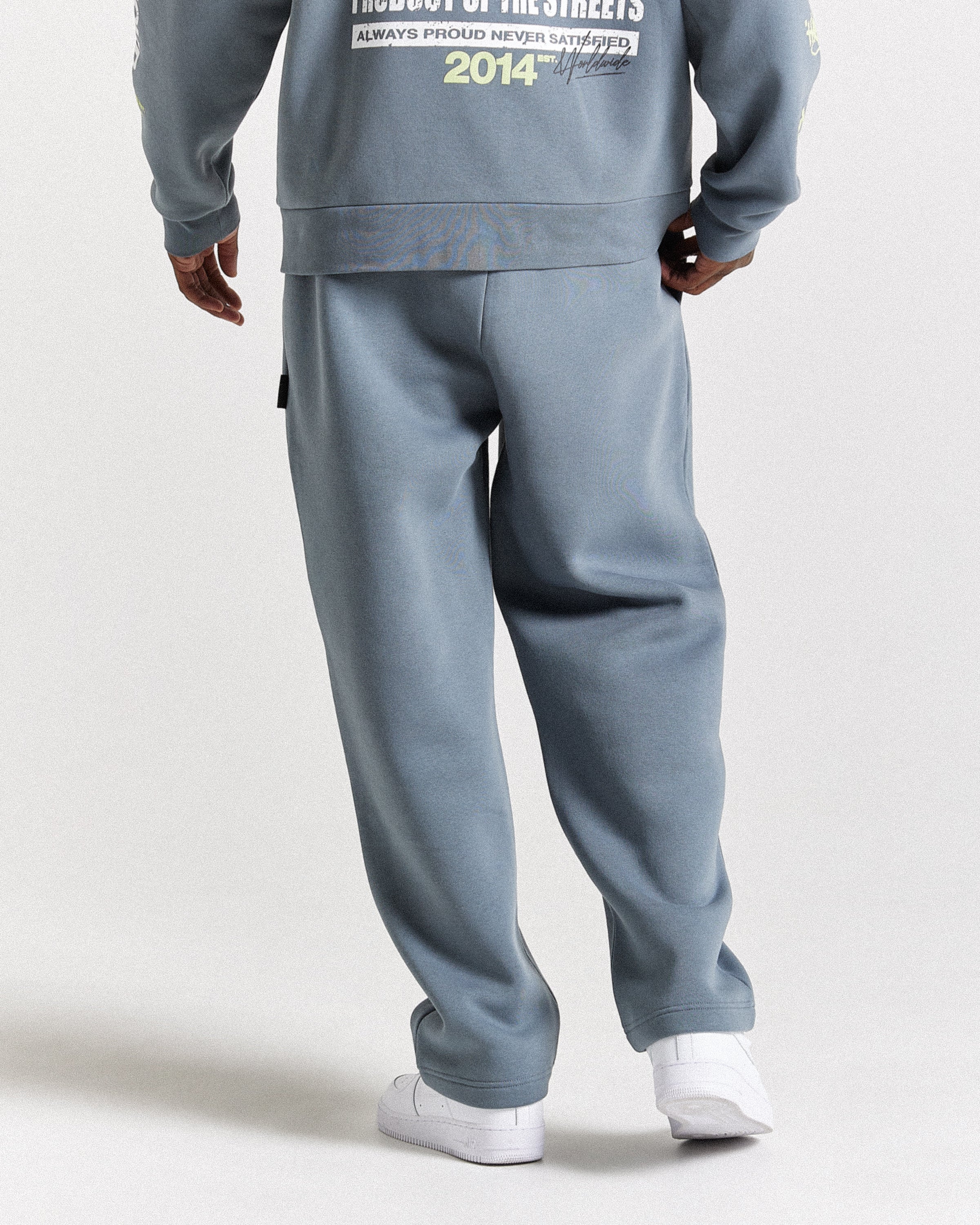A person shown from the back wearing a matching dusty blue sweatshirt and sweatpants set with white sneakers, photographed against a plain light gray studio background.