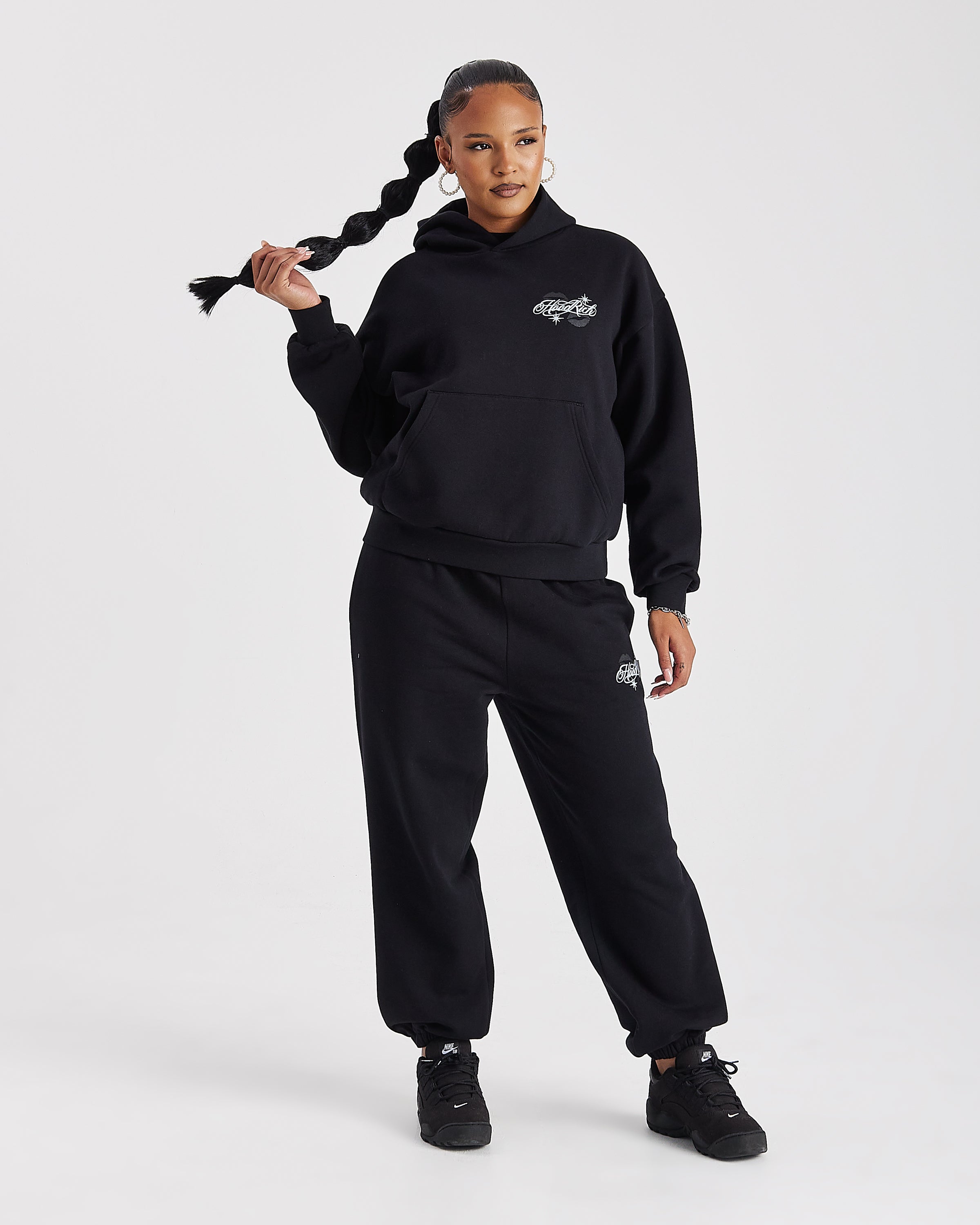 A model wearing a black hooded sweatshirt and matching sweatpants set with small white embroidered logos, posing against a light gray studio background.