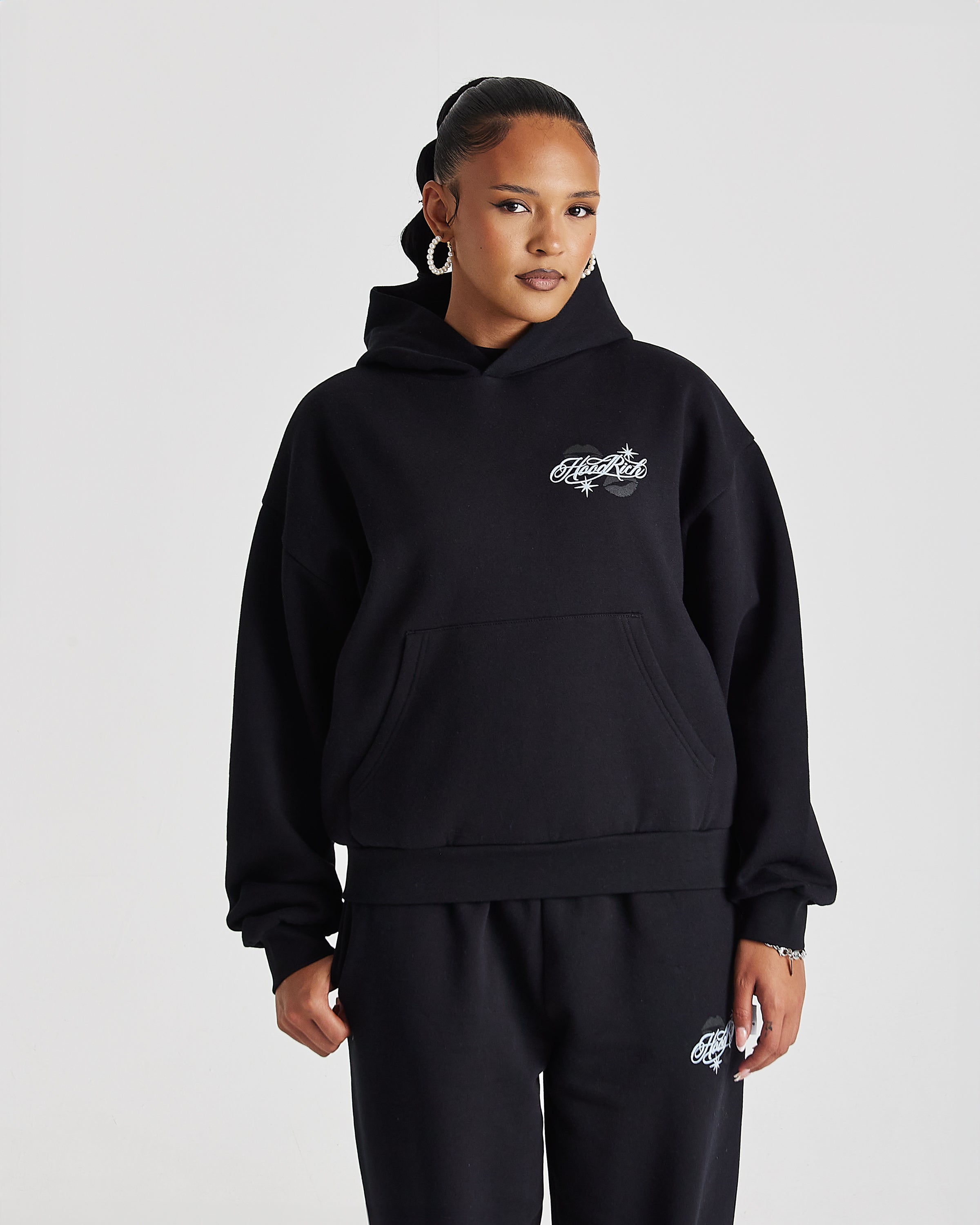 A model wearing a black pullover hoodie with a white embroidered graphic on the chest and matching black sweatpants, photographed against a light gray studio background.
