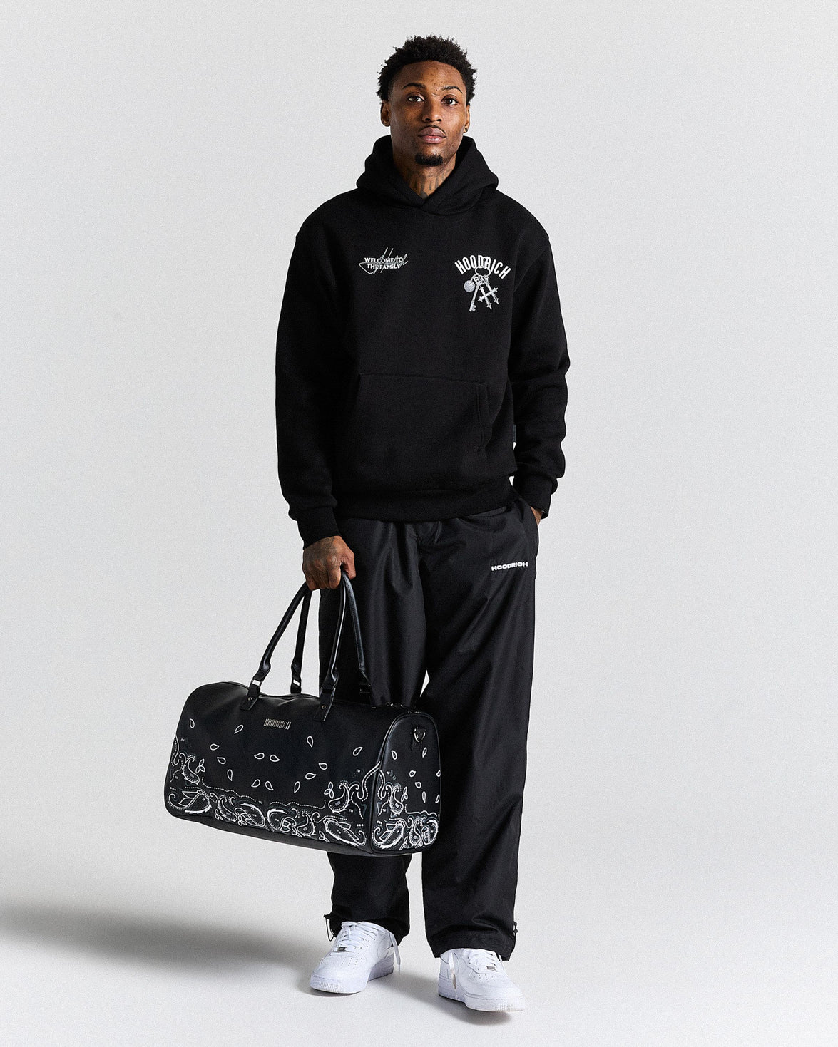 Person wearing a black hoodie and pants holding a black duffel bag with bandana design on a white background
