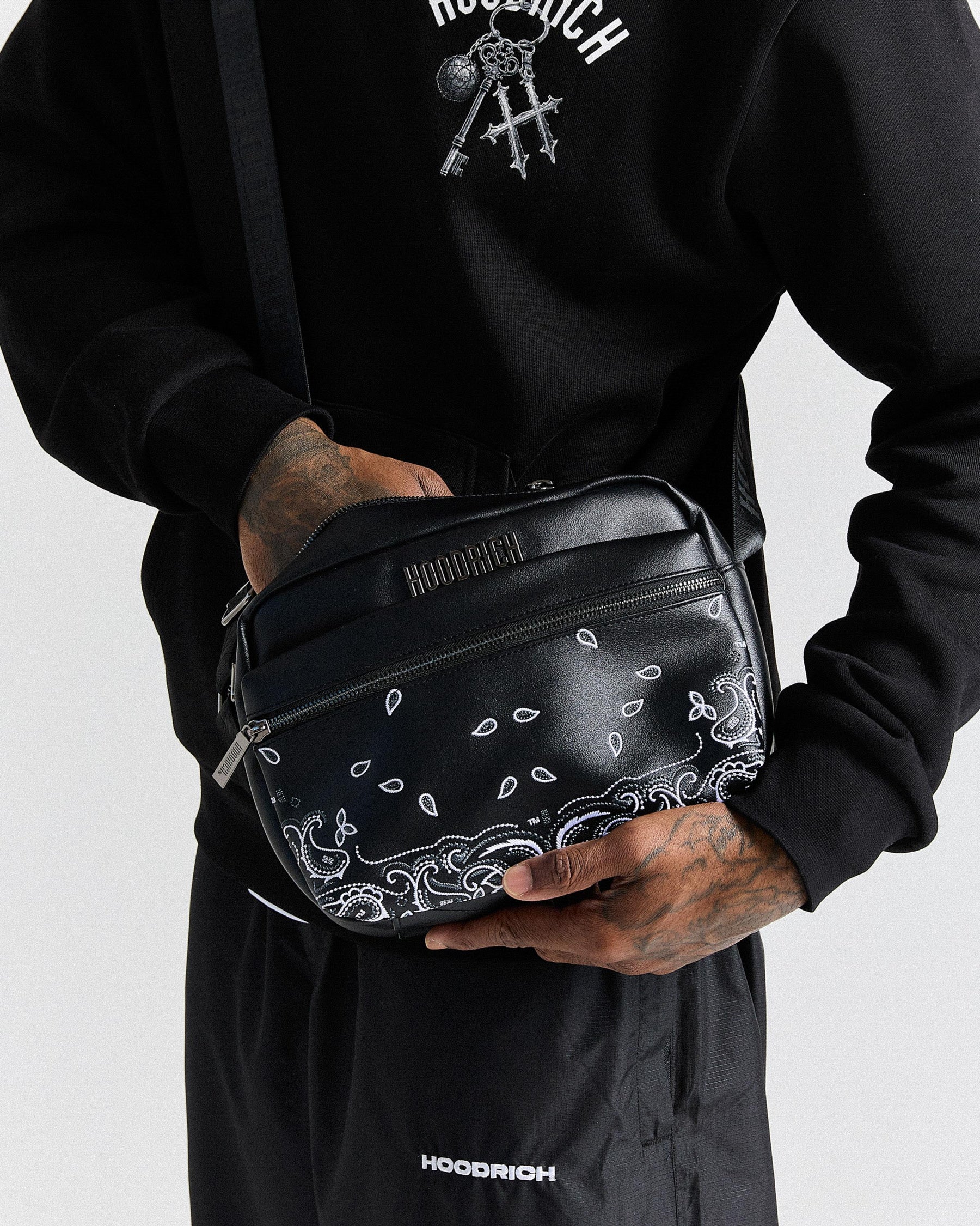 Person holding a black bag with white patterns, wearing a black hoodie and pants with 'Hoodrich' branding.
