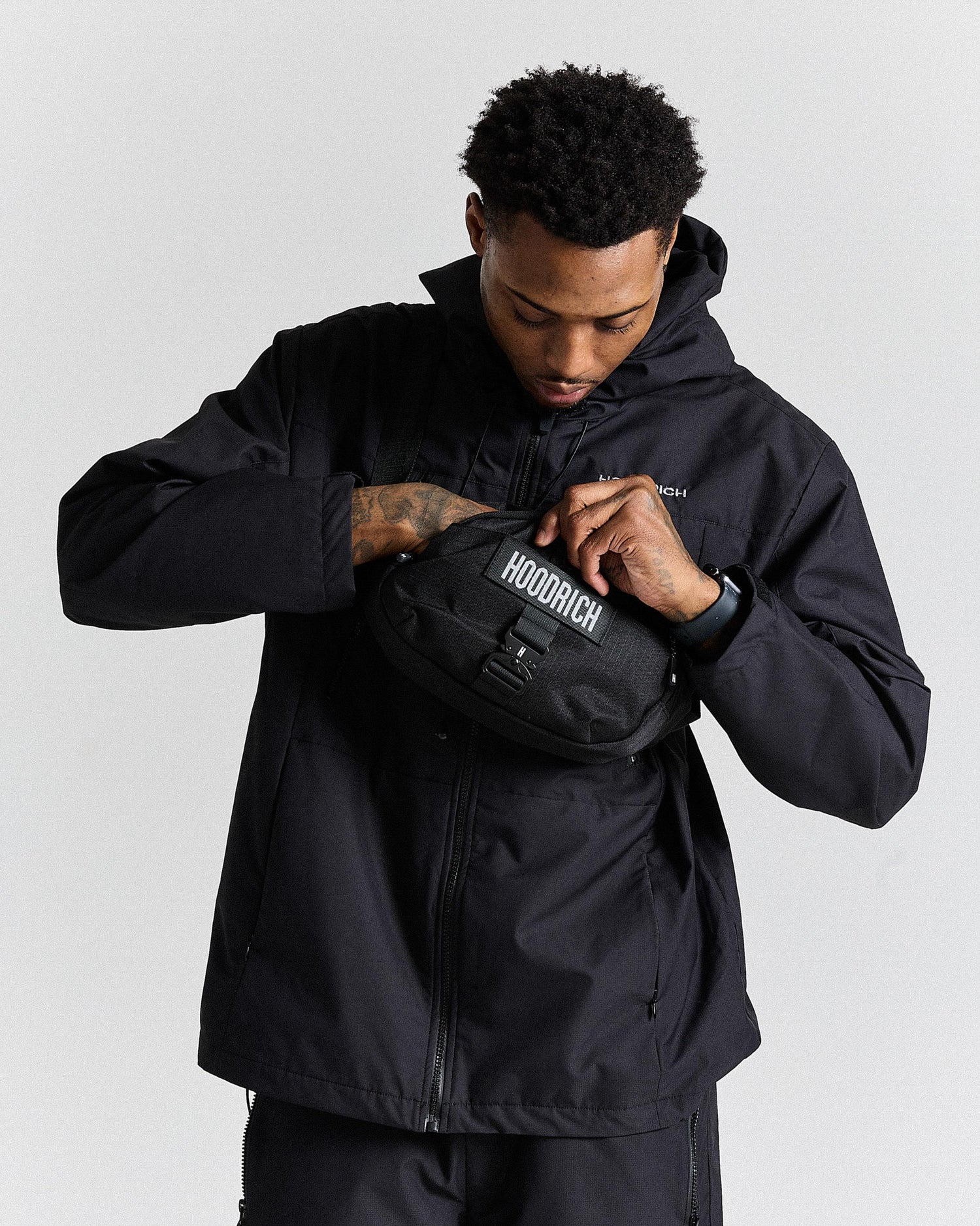 Person wearing a black jacket with a hood, holding a black bag with 'HOODRICH' branding.