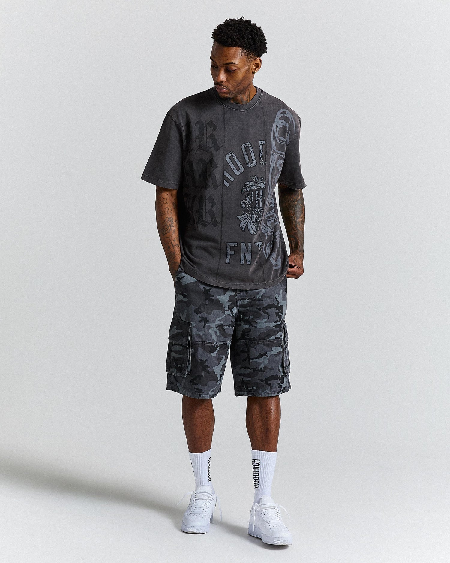 Man wearing a black graphic t-shirt and camouflage shorts on a light grey background