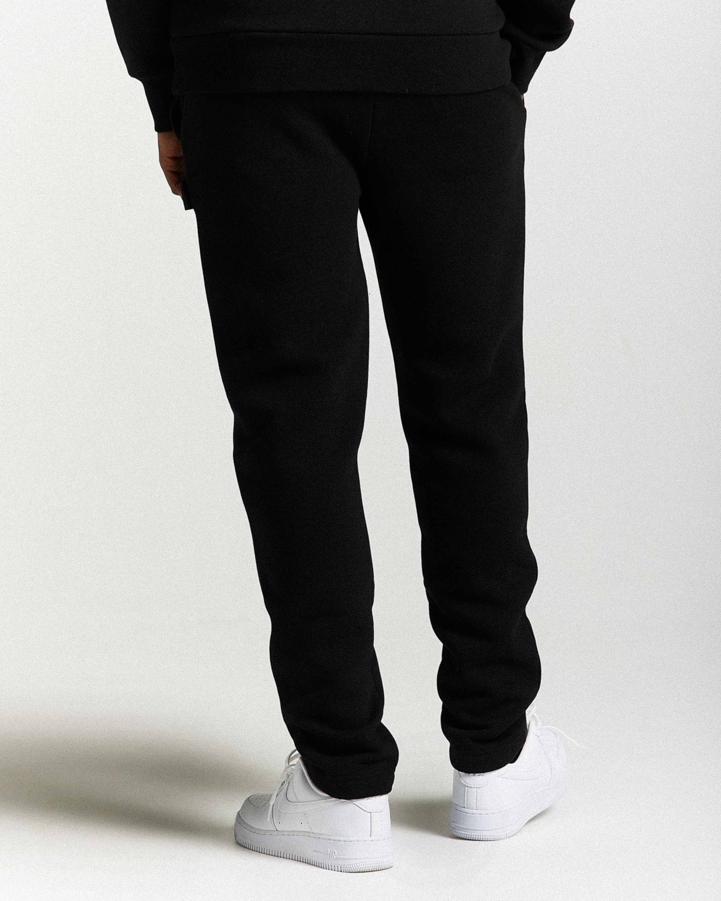 Black sweatpants shown from the back on a model, paired with white sneakers, against a plain light gray studio background.