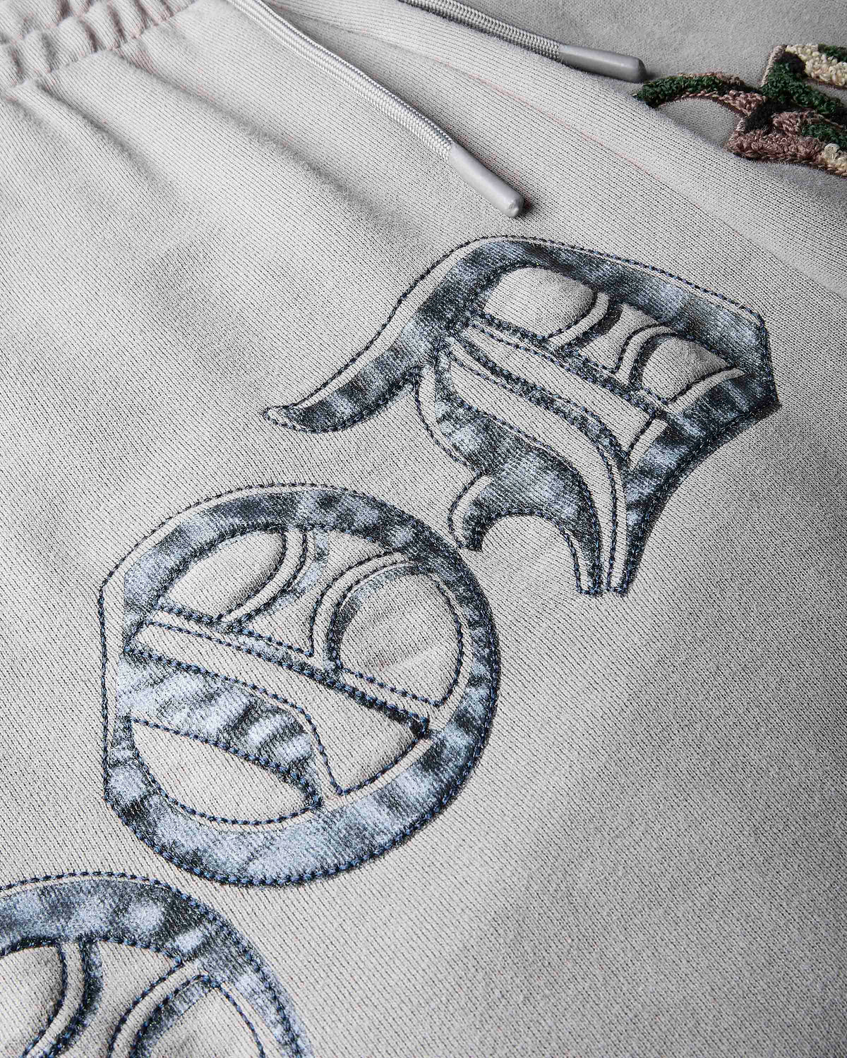 Close-up of a light beige or off-white fabric garment, likely sweatpants or a hoodie, featuring large embroidered stylized letter 'O.D' motifs in a gray and black marbled pattern. The fabric texture is highly visible, and a drawstring is visible at the top left.